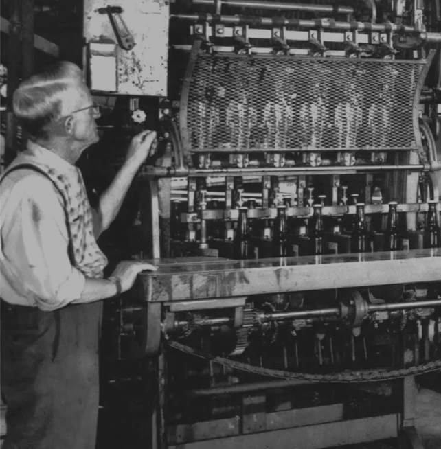 An elderly woman with glasses and two braids, wearing a plaid vest and long skirt, standing beside an early industrial machine with numerous mechanical parts, gears, and a mesh screen labeled 'GLEANER'.