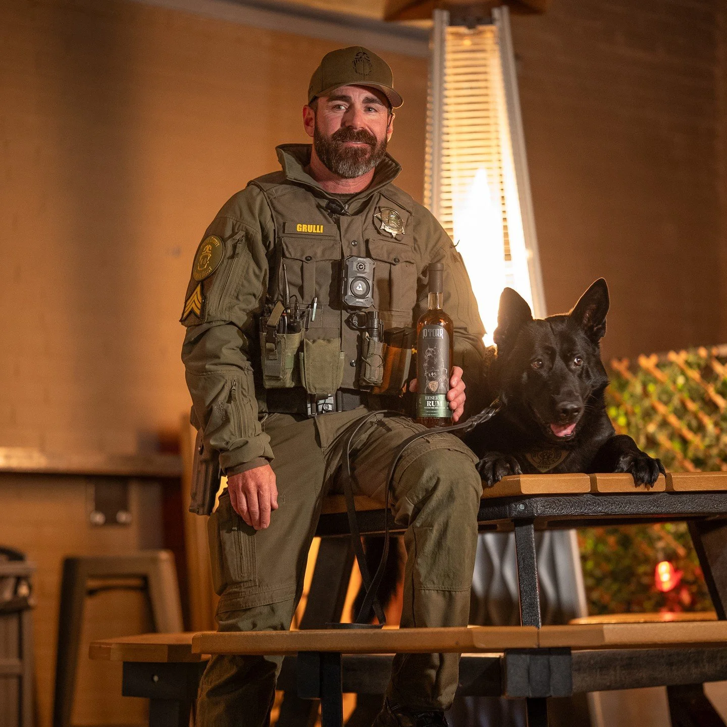 A police officer in uniform sitting on a table with a black German Shepherd police dog. The officer is holding a bottle of rum labeled "RESERVE RUM". The scene appears to be indoors with warm lighting, and there is a decorative lamp in the background.
