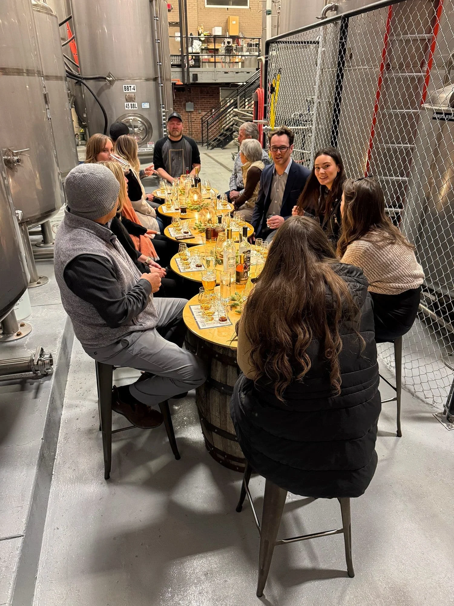 People gathered around a rustic wooden barrel table in an industrial setting, with large metal tanks and chain-link fencing in the background, enjoying drinks and conversation.