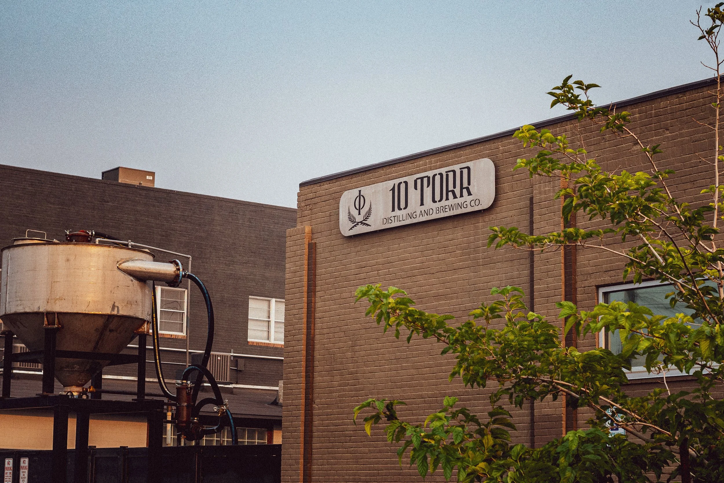 Photo of a brick building with a foam brewing company sign, trees, and rooftop equipment under a clear sky.
