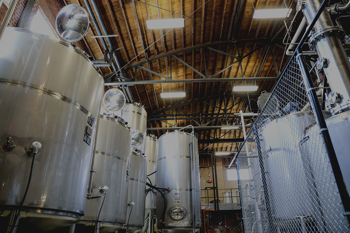Interior view of a brewery with large stainless steel fermentation tanks, wooden ceiling, and industrial lighting.