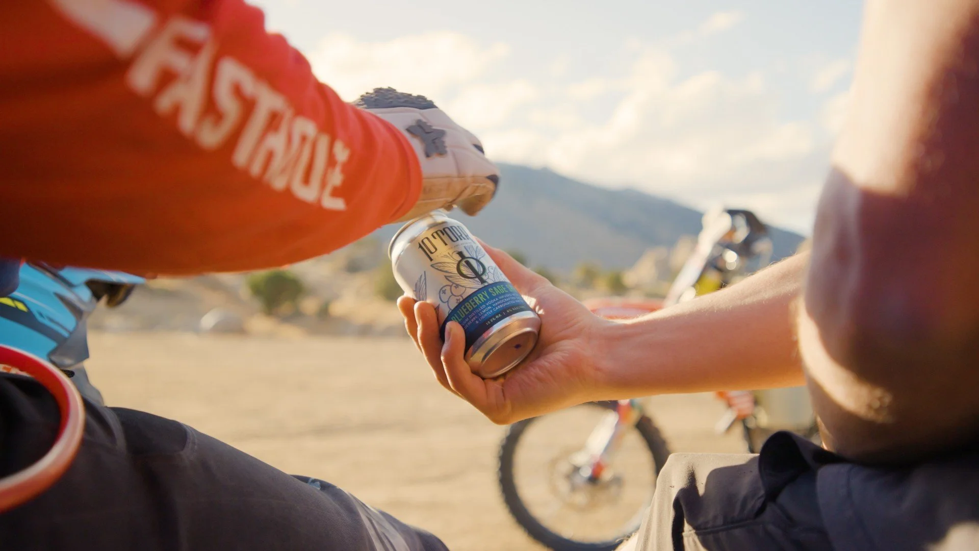 A person handing a can of blueberry sage sparkling water to another person at an outdoor location with mountains and a bicycle in the background.