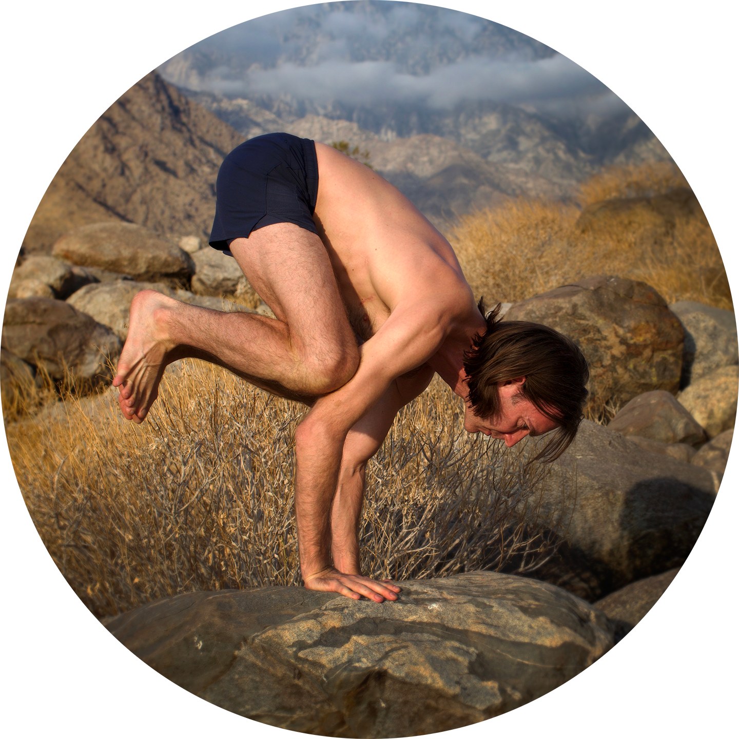 Two men practicing acro yoga outdoors on rocks and dry grass, with mountains and cloudy sky in the background.