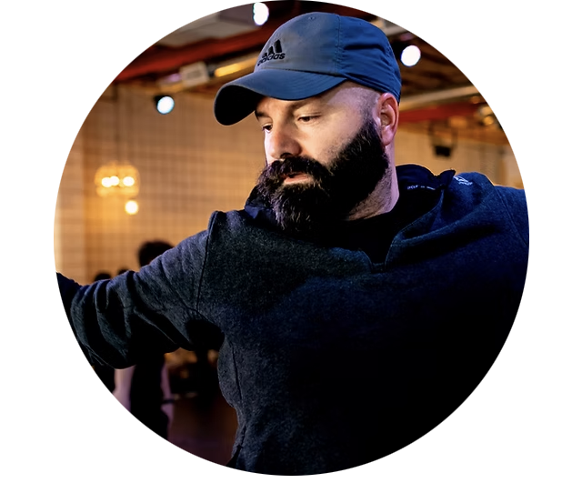 A bearded man wearing a blue baseball cap and a dark hoodie, looking down in a dimly lit indoor setting.
