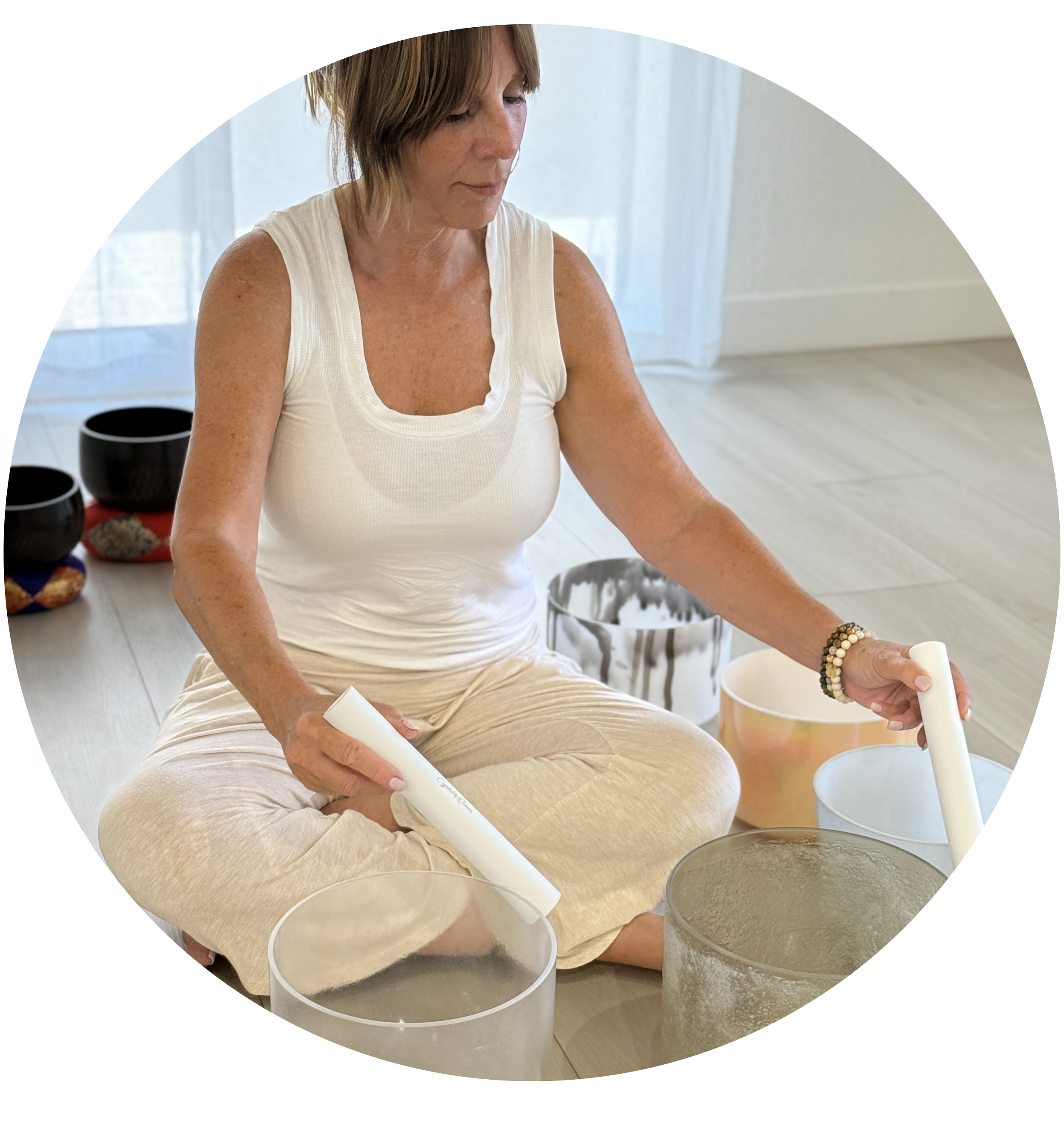 A woman with short reddish hair and freckles, wearing a white sleeveless top and beige pants, is sitting cross-legged on the floor, rolling a white candle with both hands. She has beaded bracelets on her right wrist. In front of her are multiple large white candles and small black singing bowls behind her. The setting appears to be bright, with sheer white curtains and a light-colored floor in the background.