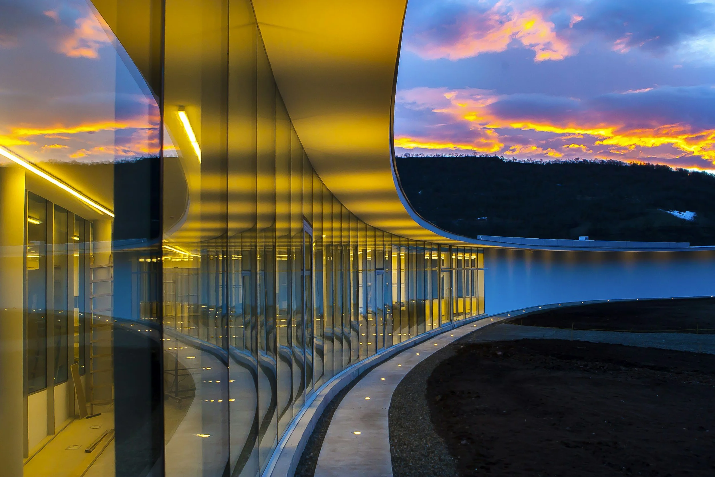 A modern building with large glass windows and a curved yellow roof at sunset, reflecting the colorful sky in the glass, with mountains and a partly cloudy sky in the background.
