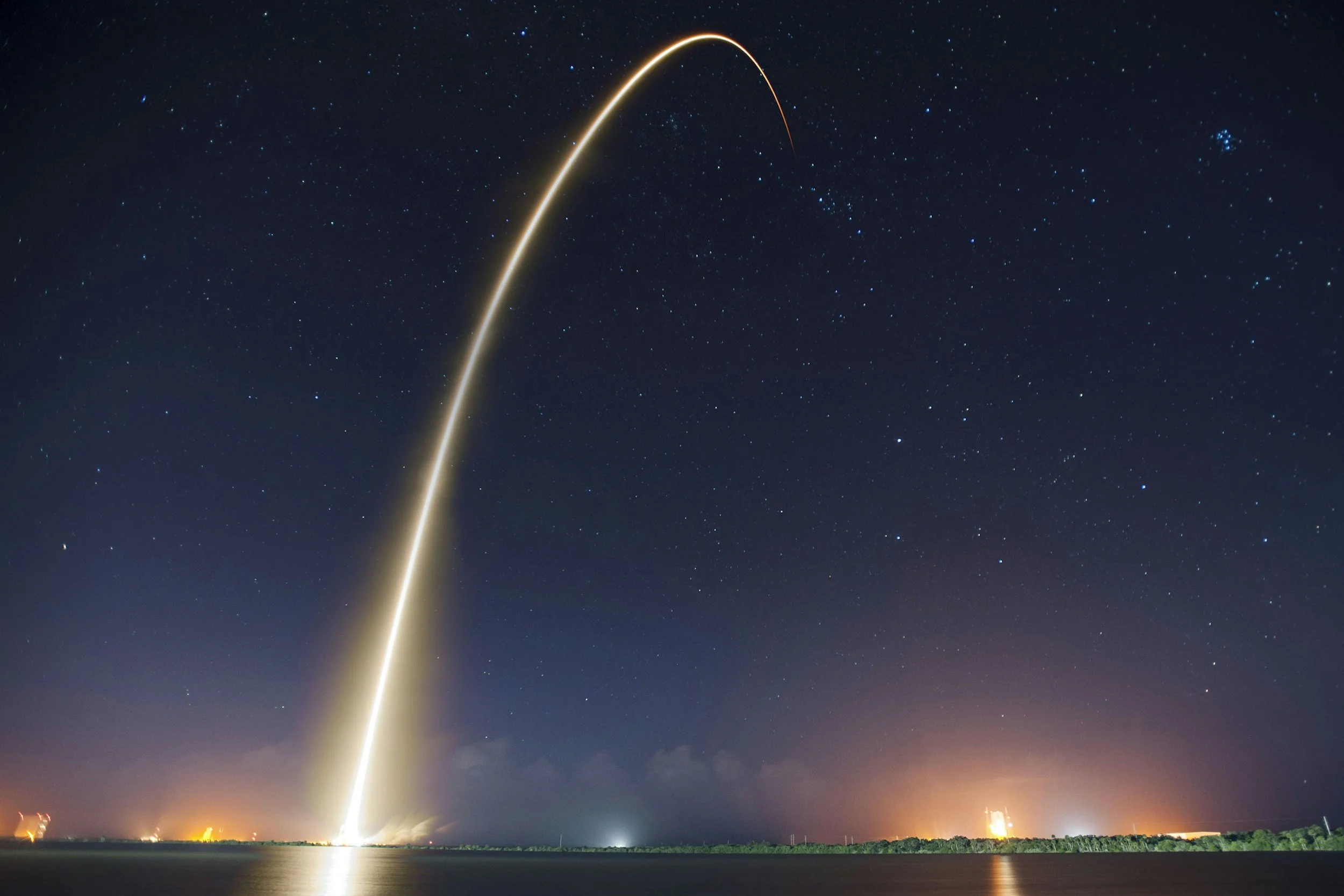A rocket launch at night with a trail of bright light against a starry sky over a body of water.
