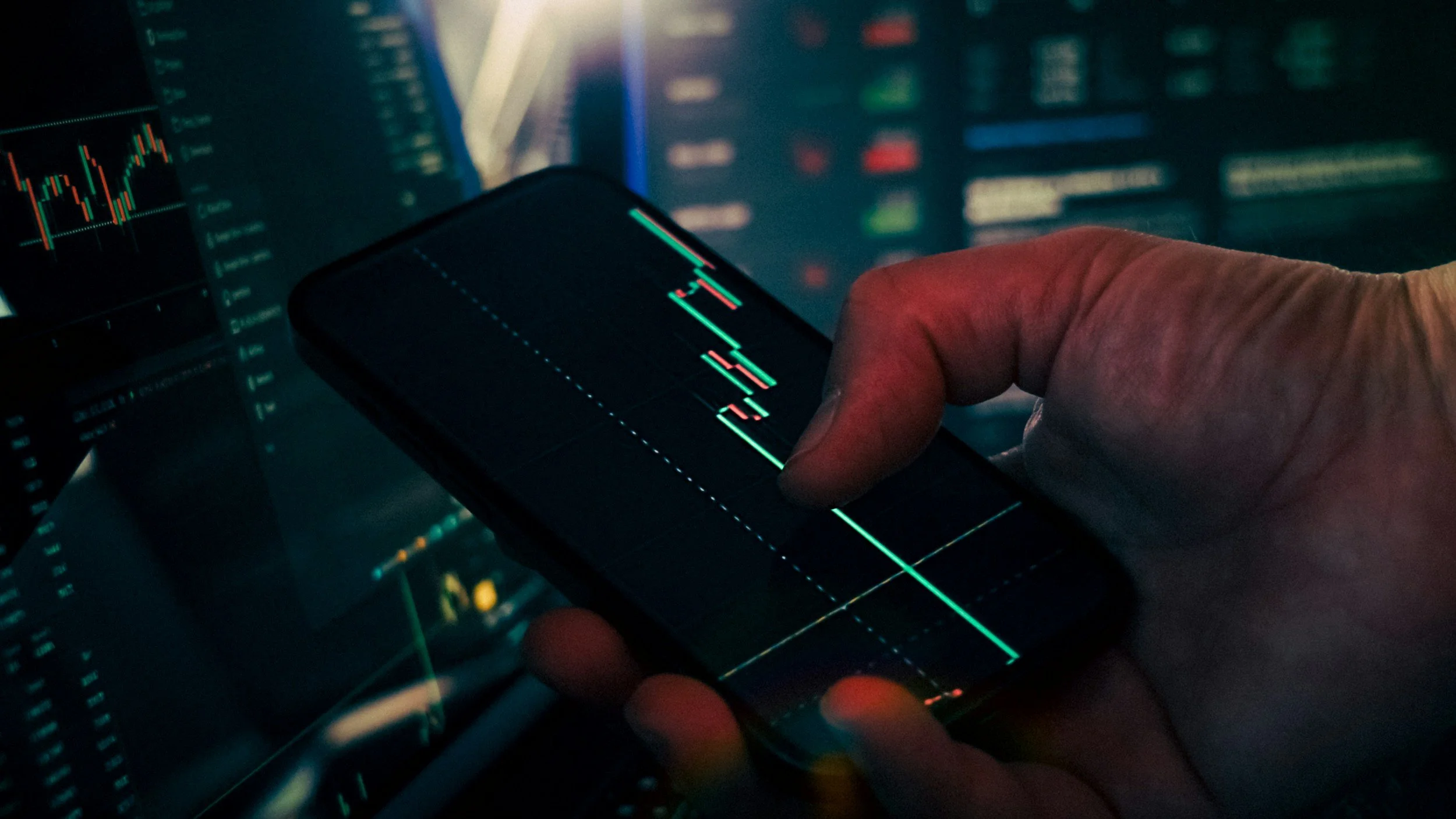 Close-up of a person's hand holding a smartphone displaying a stock market chart with red and green candlesticks, with computer screens in the background showing financial data and graphs.