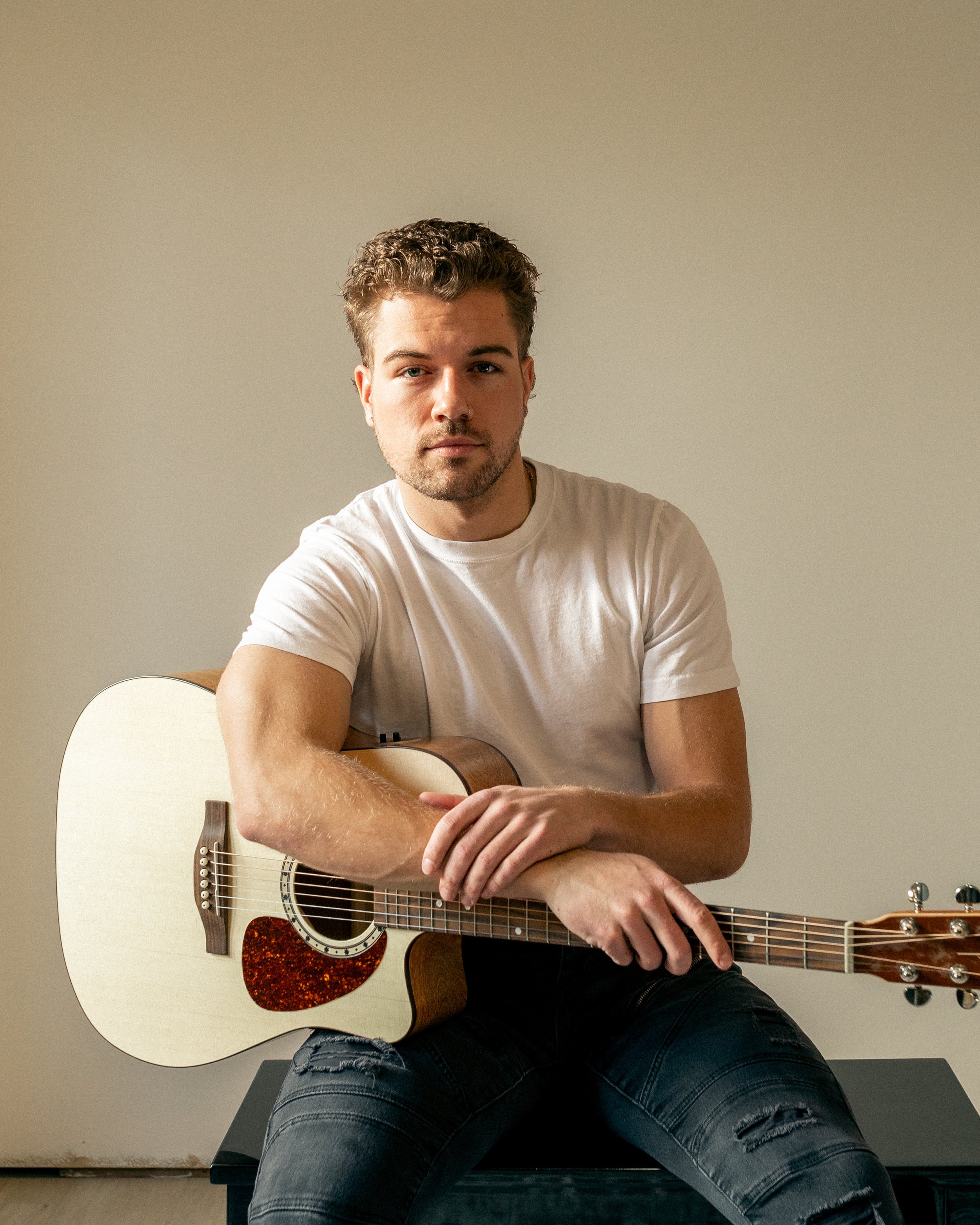 Matt Johner. A young man with curly hair and a beard sitting on a black ottoman, holding an acoustic guitar across his lap, wearing a white T-shirt and ripped jeans, against a plain beige wall.