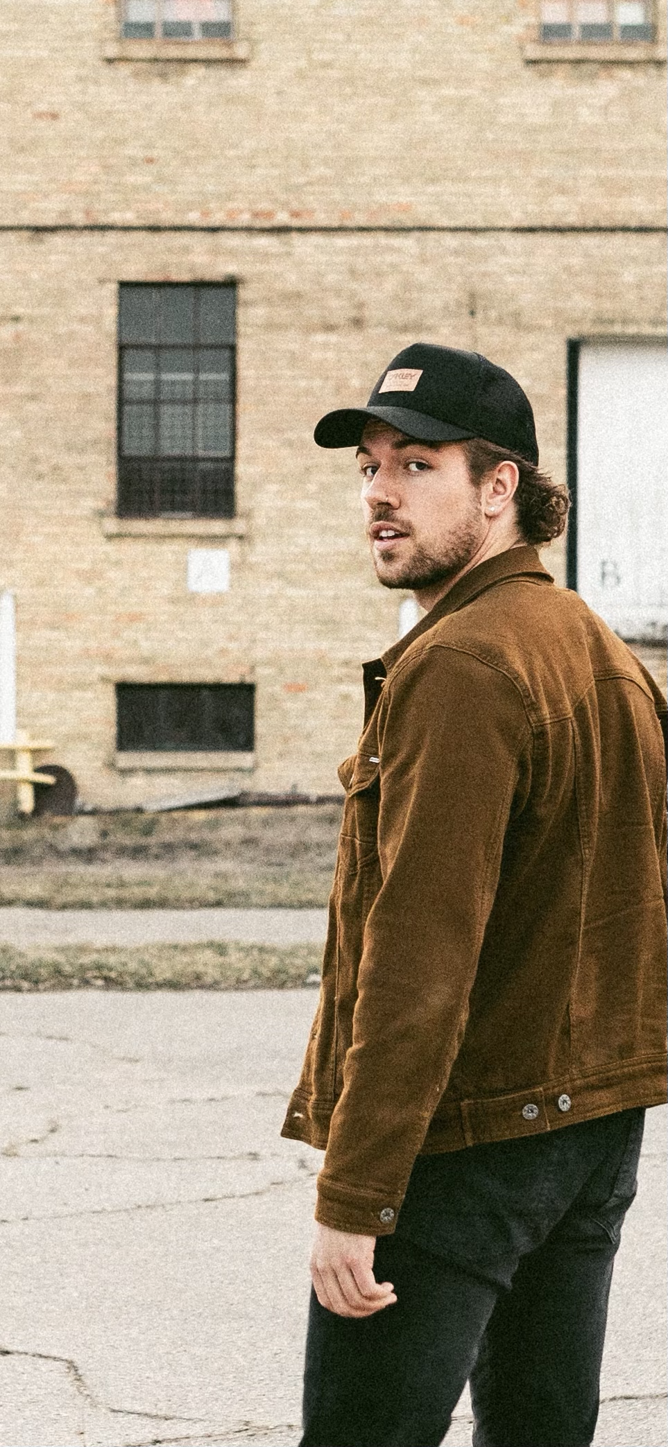 A young man with curly hair and a beard, wearing a black cap and brown jacket, standing outdoors on a street with a brick building in the background.