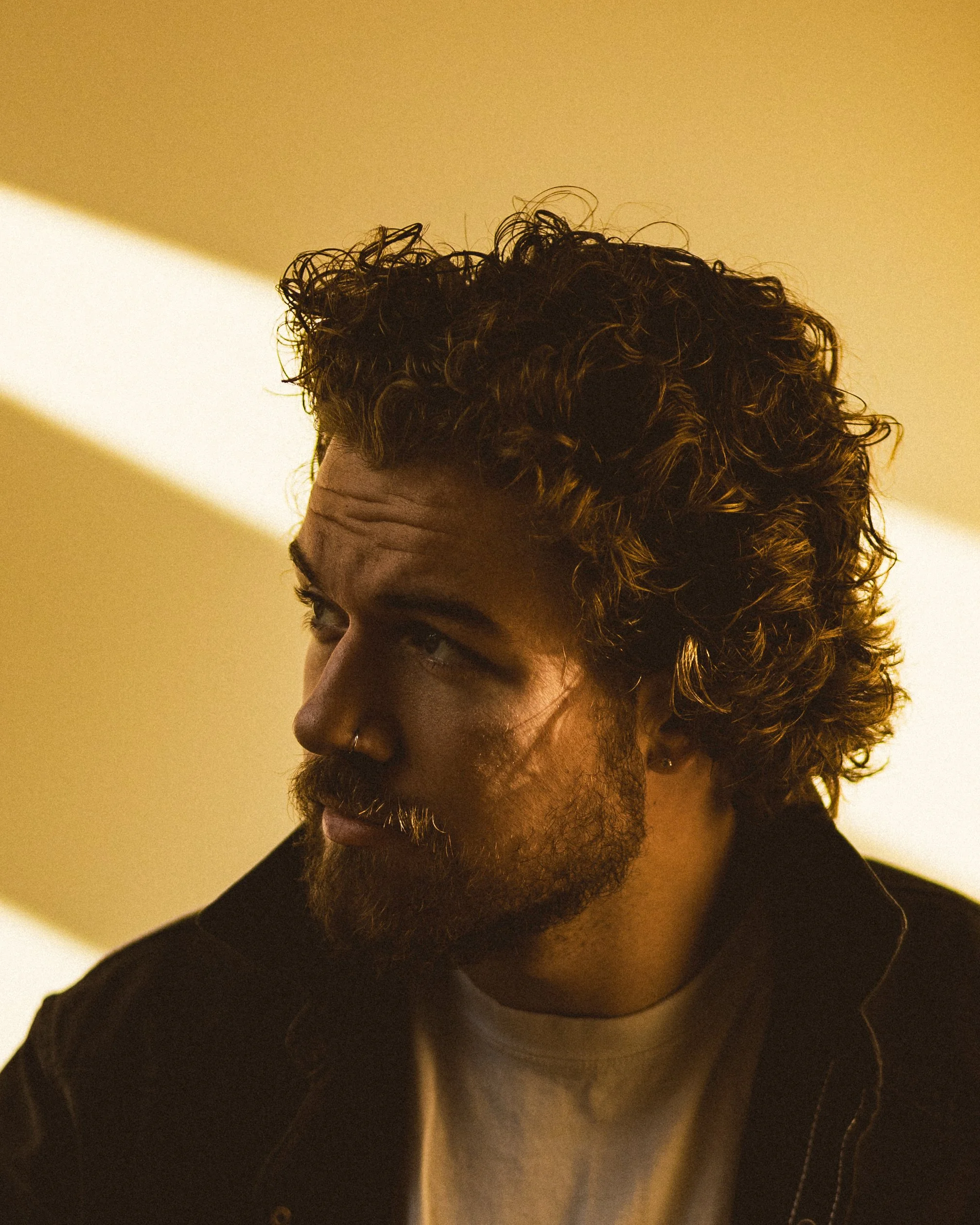 Matt Johner. A close-up of a man's face and curly hair, with a beige background and shadows.