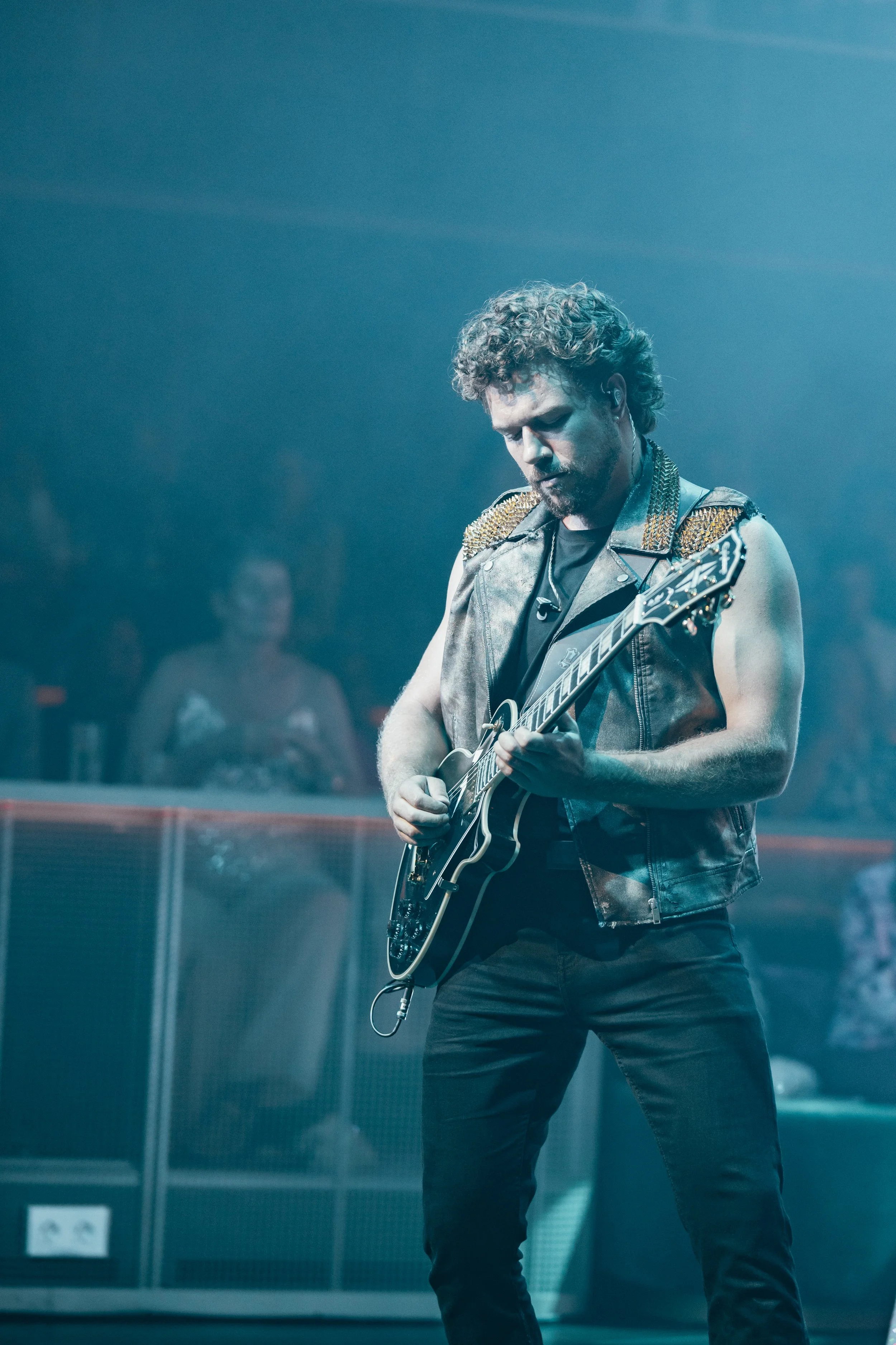 A male musician playing an electric guitar on stage during a concert, wearing a sleeveless leather vest with decorative studs.