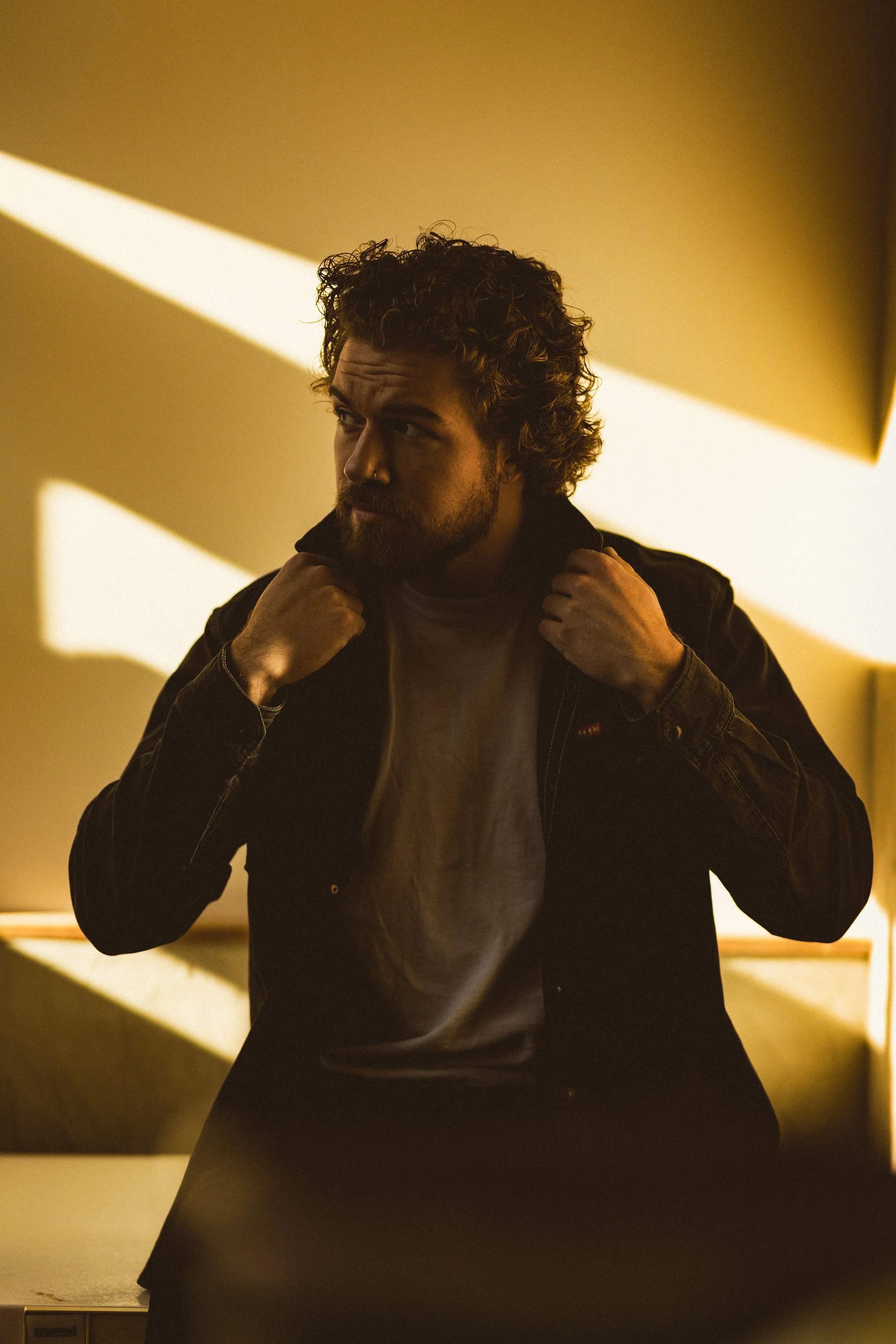 A man with curly hair and a beard is sitting in a dimly lit room with warm yellow lighting. He is wearing a dark jacket and appears to be adjusting his collar while looking to the side. The background features geometric shadows cast by the lighting.