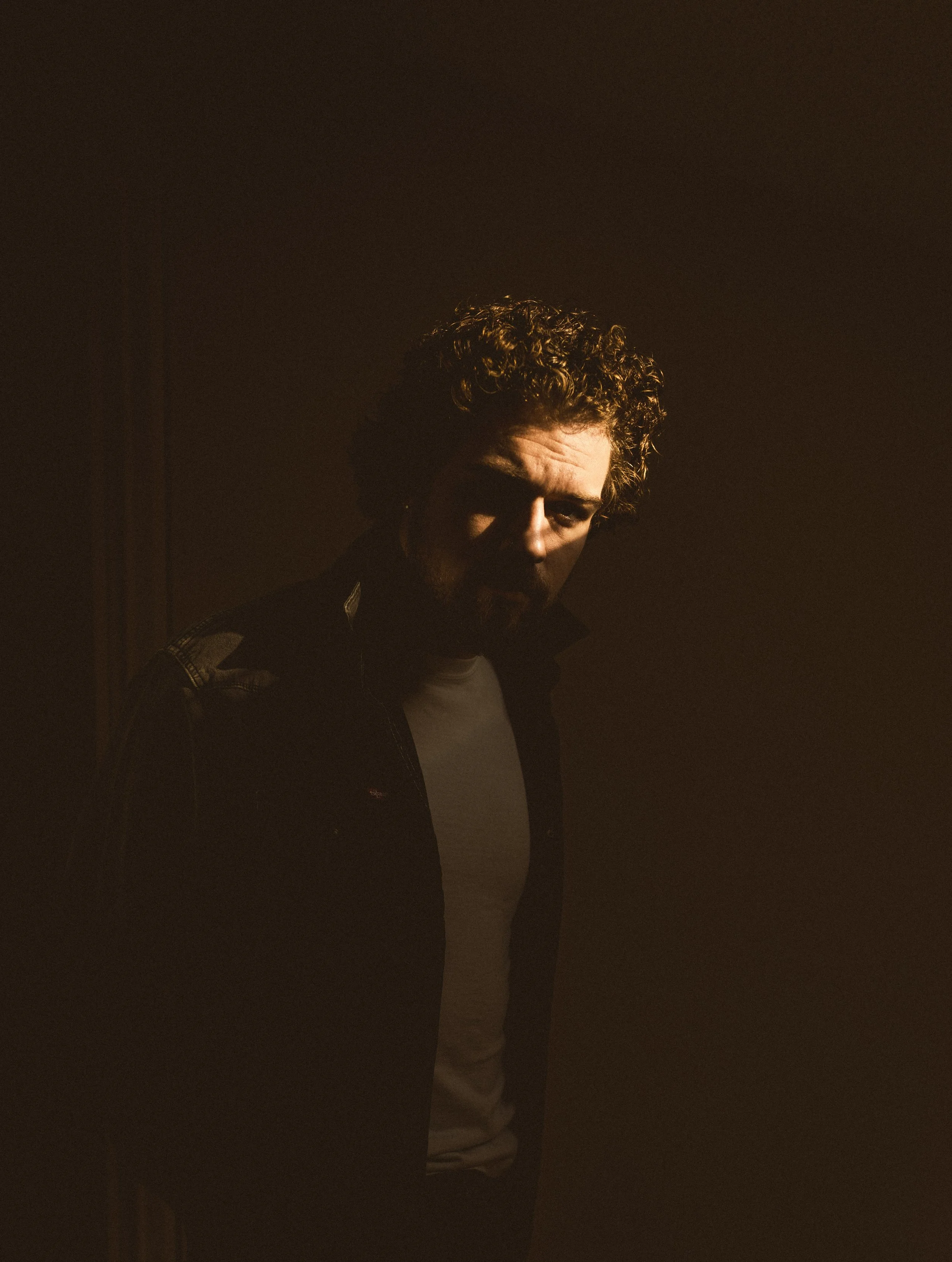 Matt Johner. A man with curly hair and a beard looking down, illuminated softly from the front, with a dark background.