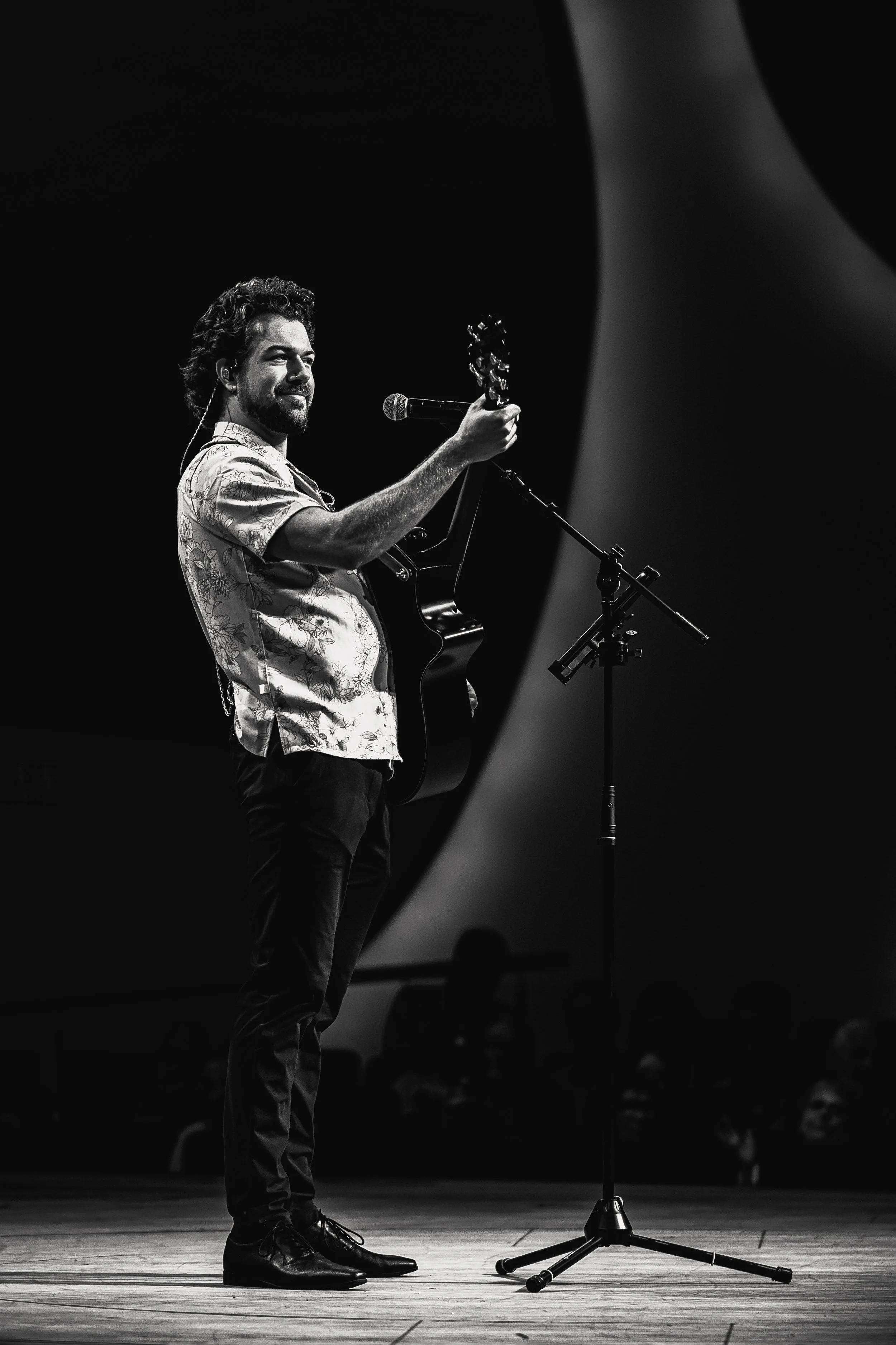A man with curly hair and a beard stands on a wooden stage, holding a guitar in his right hand. He is wearing a patterned short-sleeve shirt and dark pants, smiling slightly. In front of him is a microphone on a stand, and the background is dark with a spotlight illuminating him and part of the stage floor.