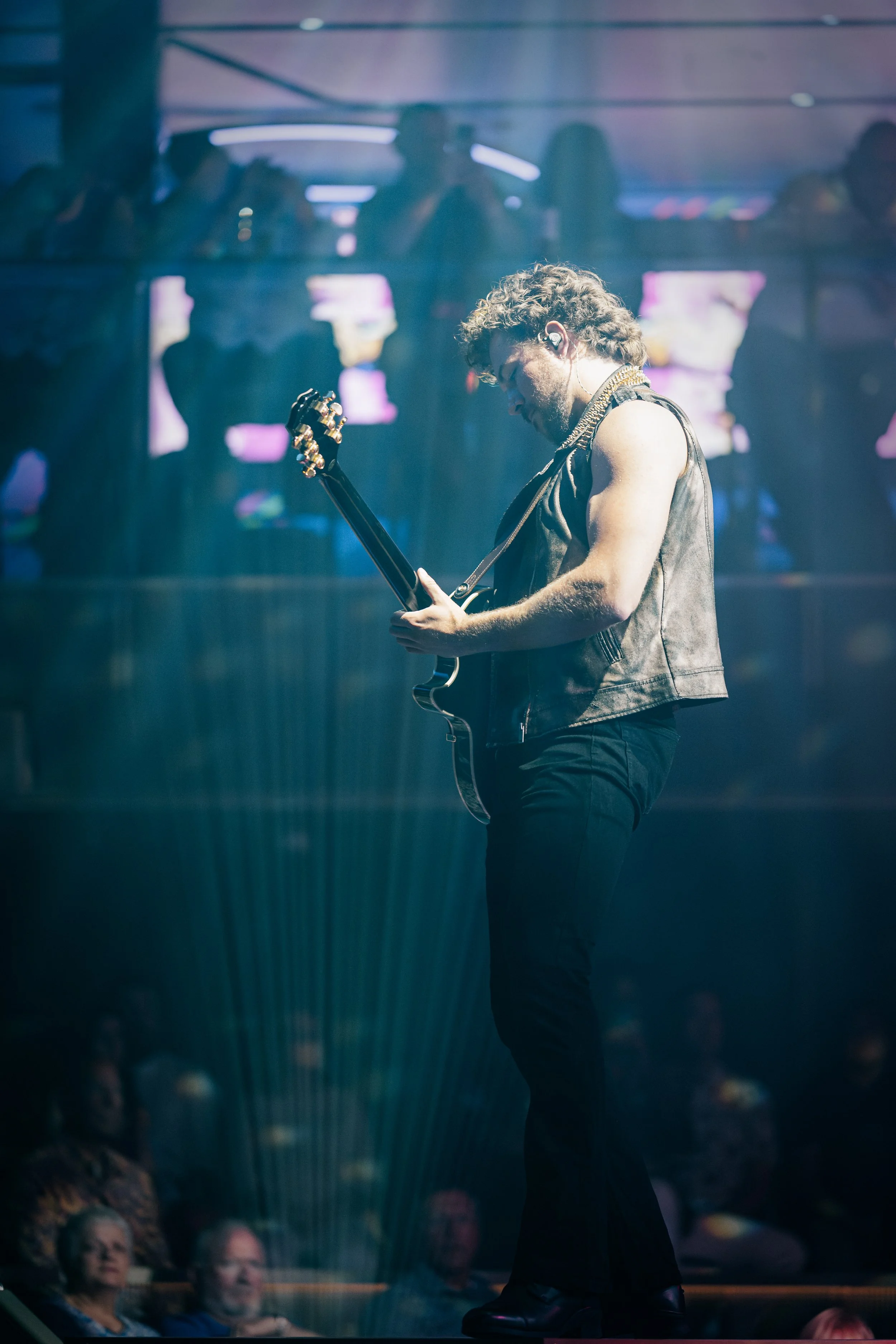 A young man with curly hair and a beard playing an electric guitar on stage. He is wearing a sleeveless leather vest and black pants, with a headset microphone. The background shows an audience watching him perform, with some audience members visible