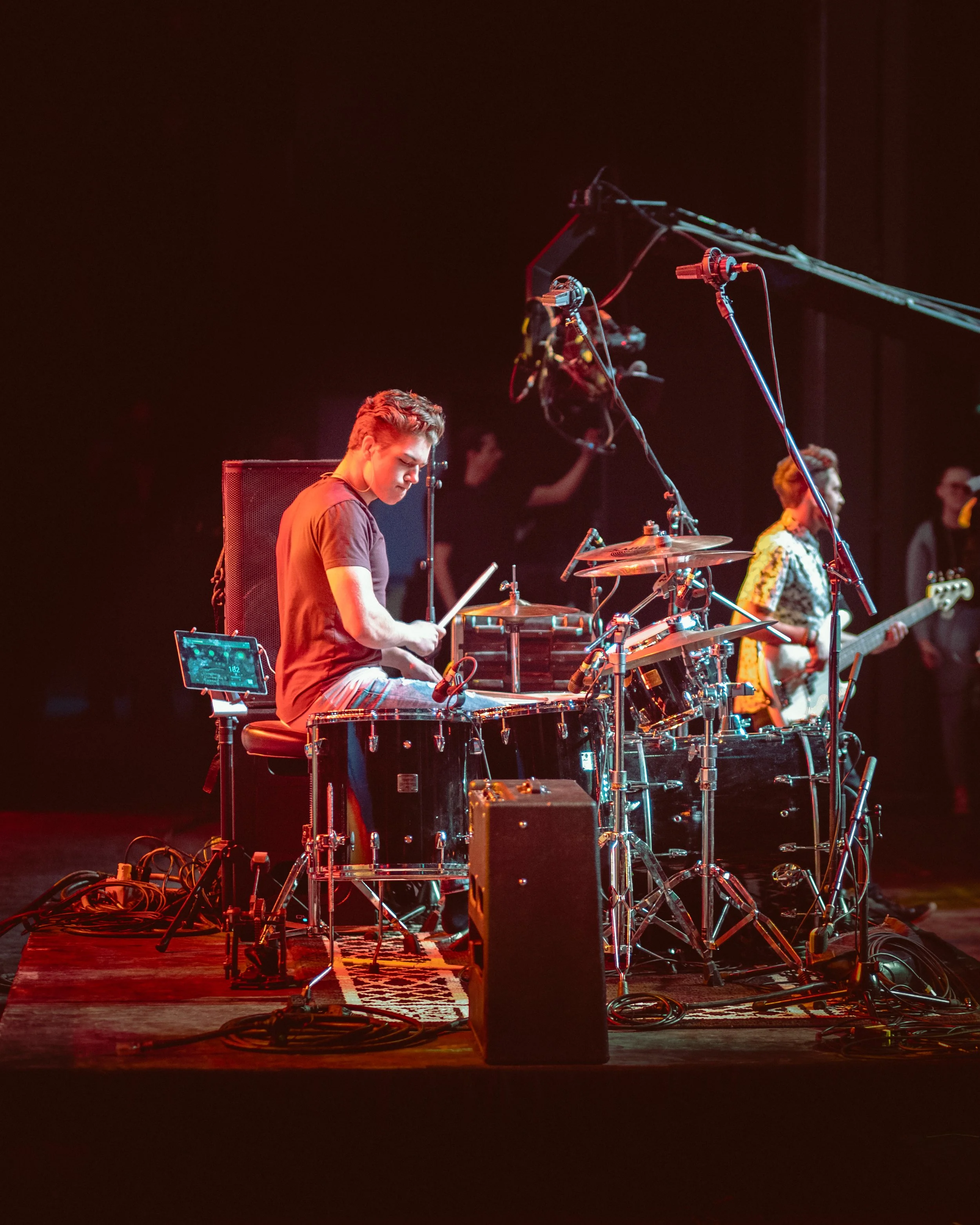 Matt Johner. A musician playing drums on stage with other band members, microphones, and stage equipment visible, dark background with stage lighting.
