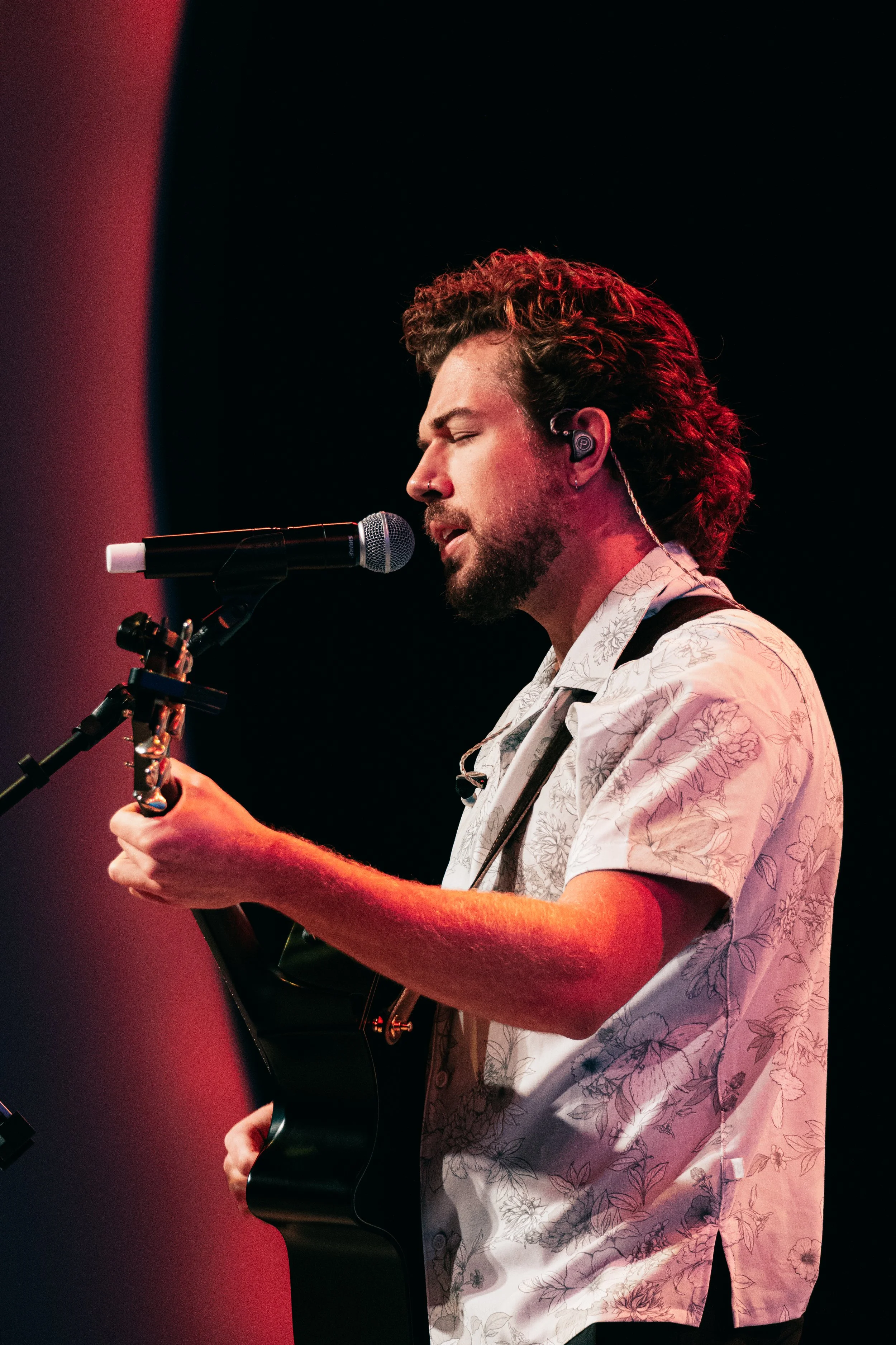 A man with curly brown hair and a beard passionately singing into a microphone while playing an acoustic guitar, wearing a white floral shirt, with a dark background and red lighting.