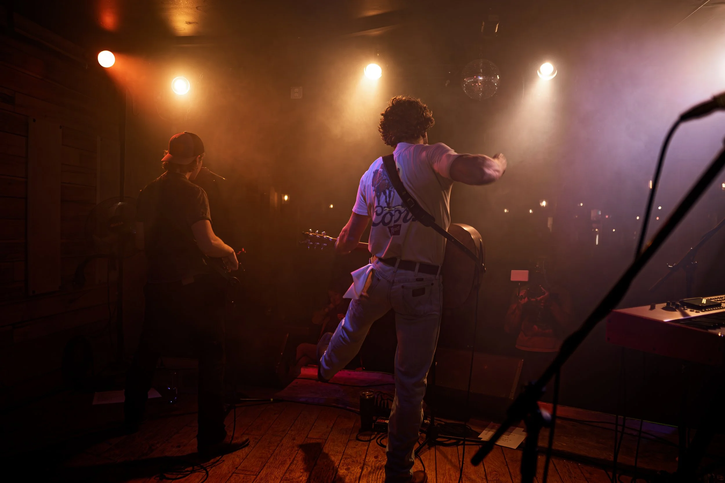 Two musicians performing on a dimly lit stage with warm, amber lighting. One is playing the guitar and the other is singing into a microphone. A discoball and some audio equipment are visible. The atmosphere is intimate and moody.