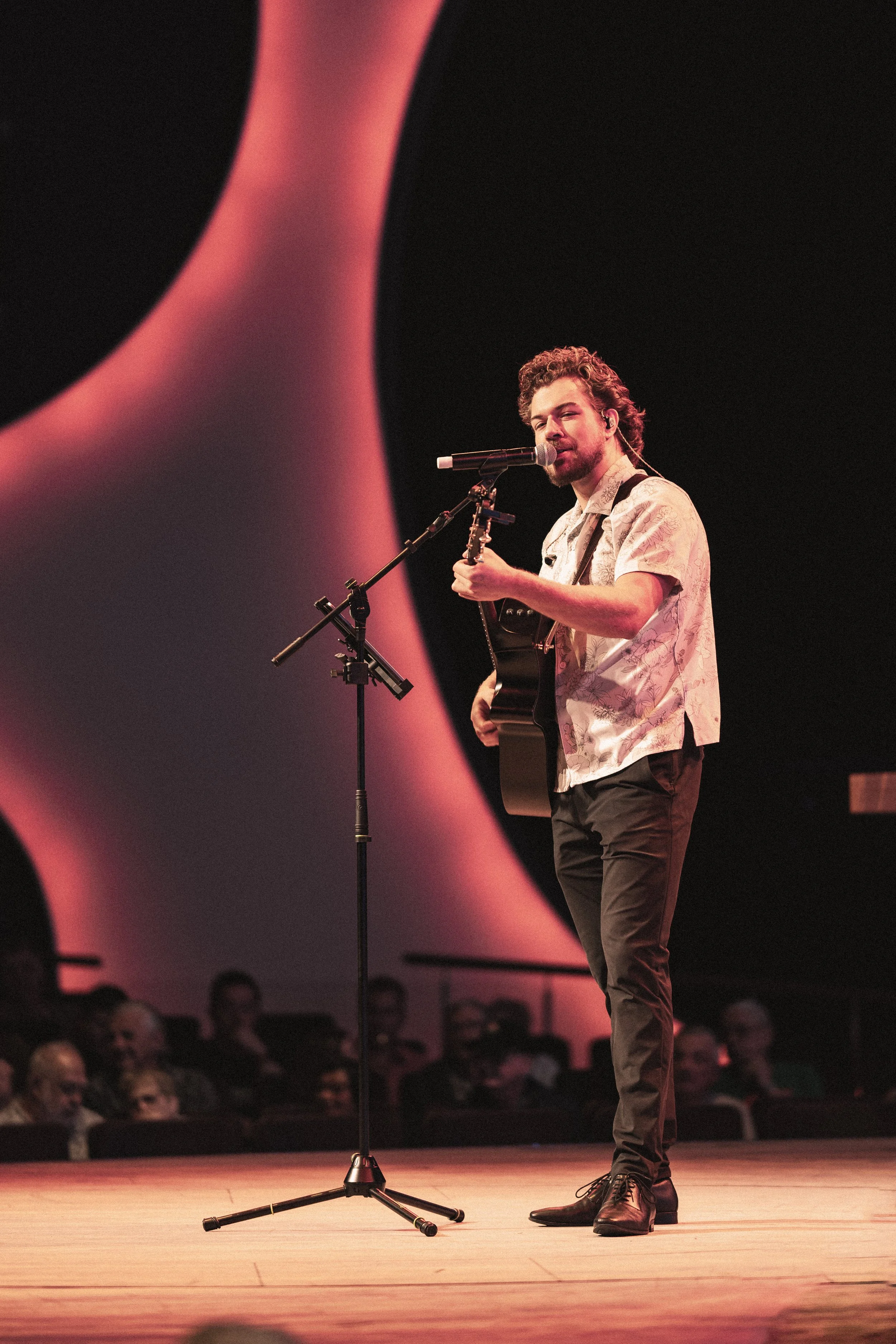 A male singer with curly hair and beard performs on stage, playing a guitar and singing into a microphone. Audience members are seated in the background.