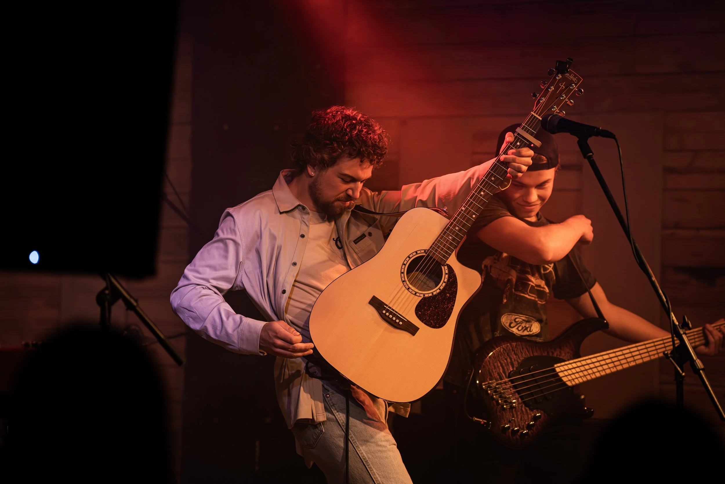 Two young men performing on stage, one playing an acoustic guitar and the other holding an electric guitar, in a dimly lit venue with wooden wall backdrop.