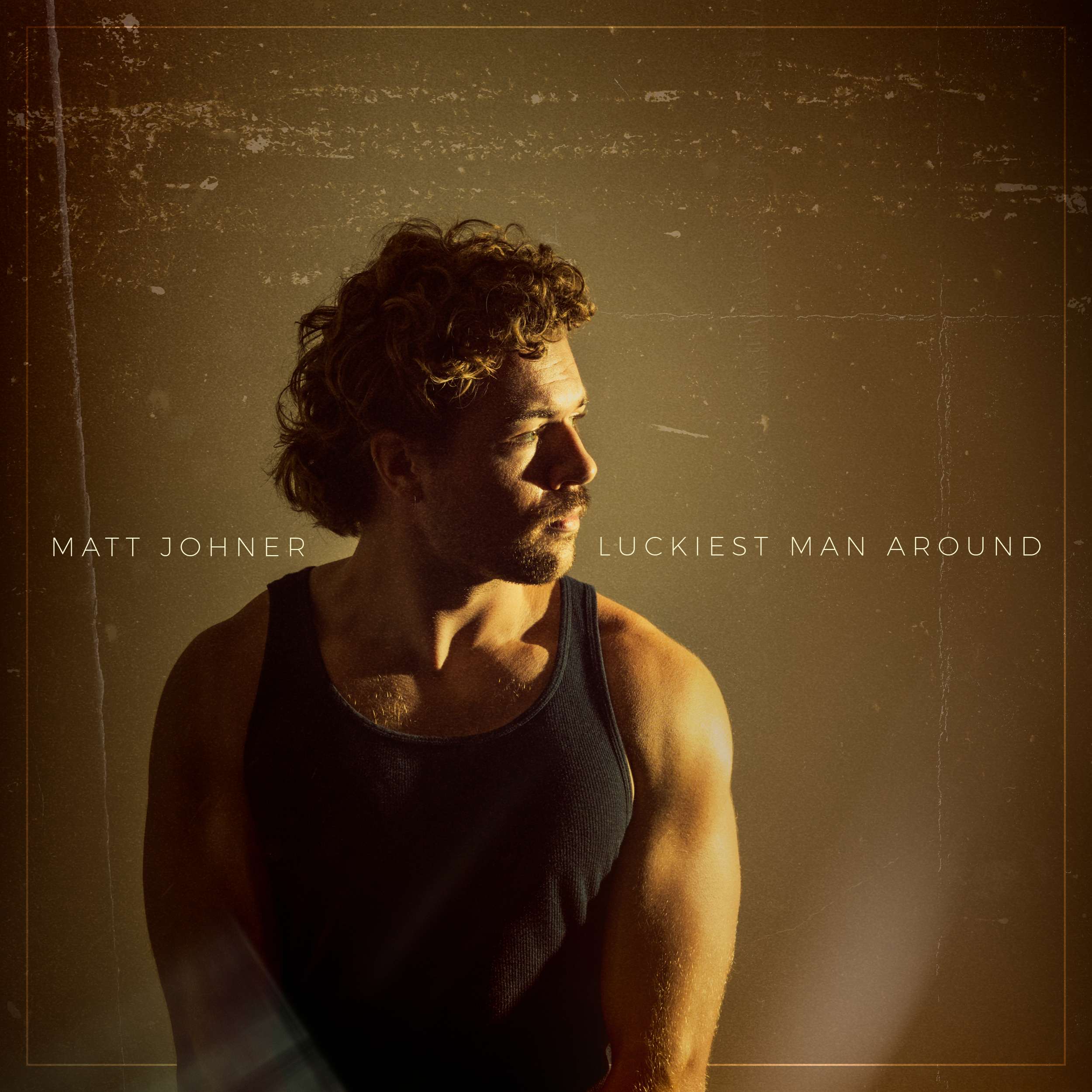 Matt Johner. Side profile of a man with curly hair and beard, wearing a black tank top, sunlit against a dark textured background.