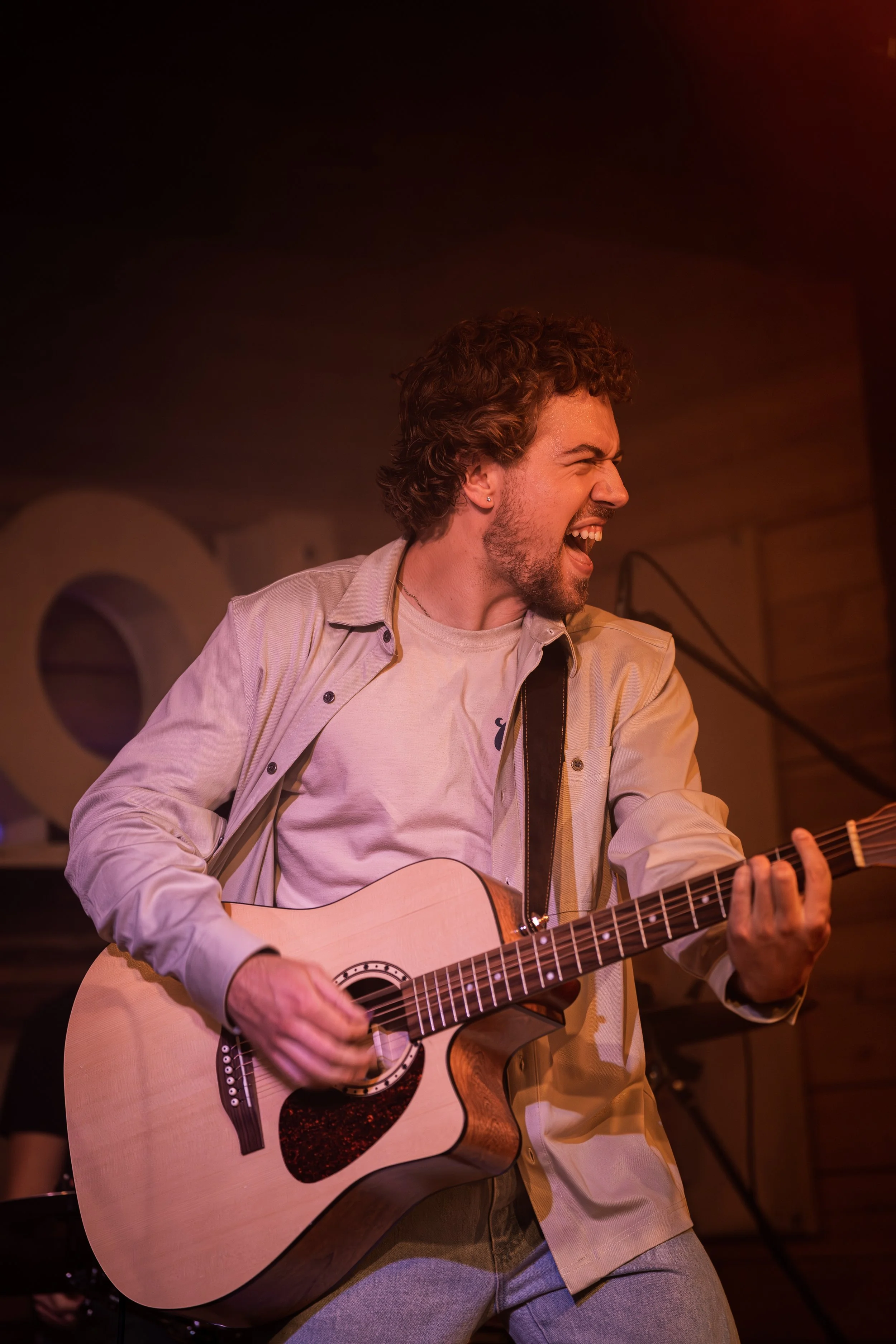 A young man with curly hair and a beard passionately singing and playing an acoustic guitar on stage.