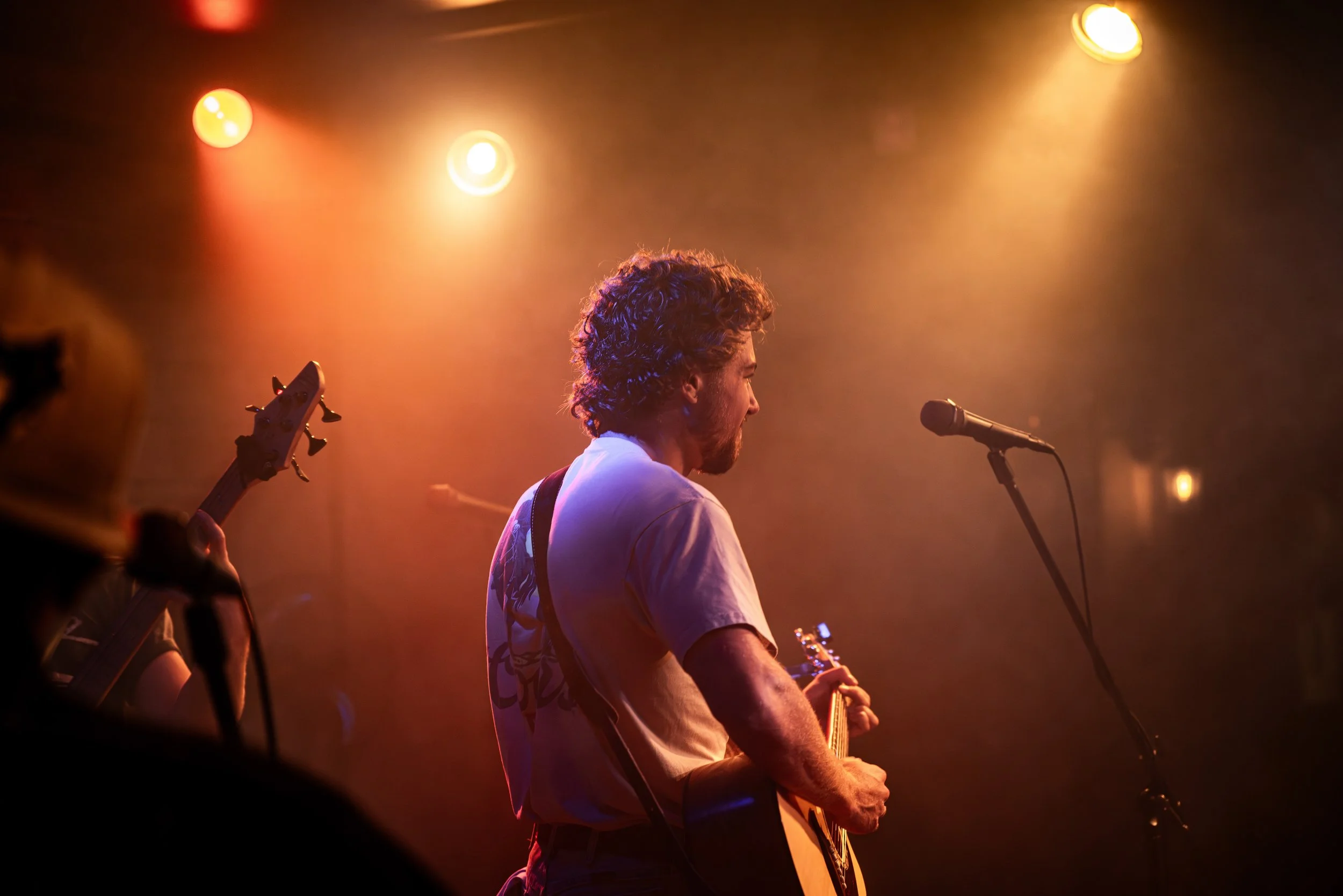 A man playing guitar on stage with stage lights overhead and a microphone in front of him.