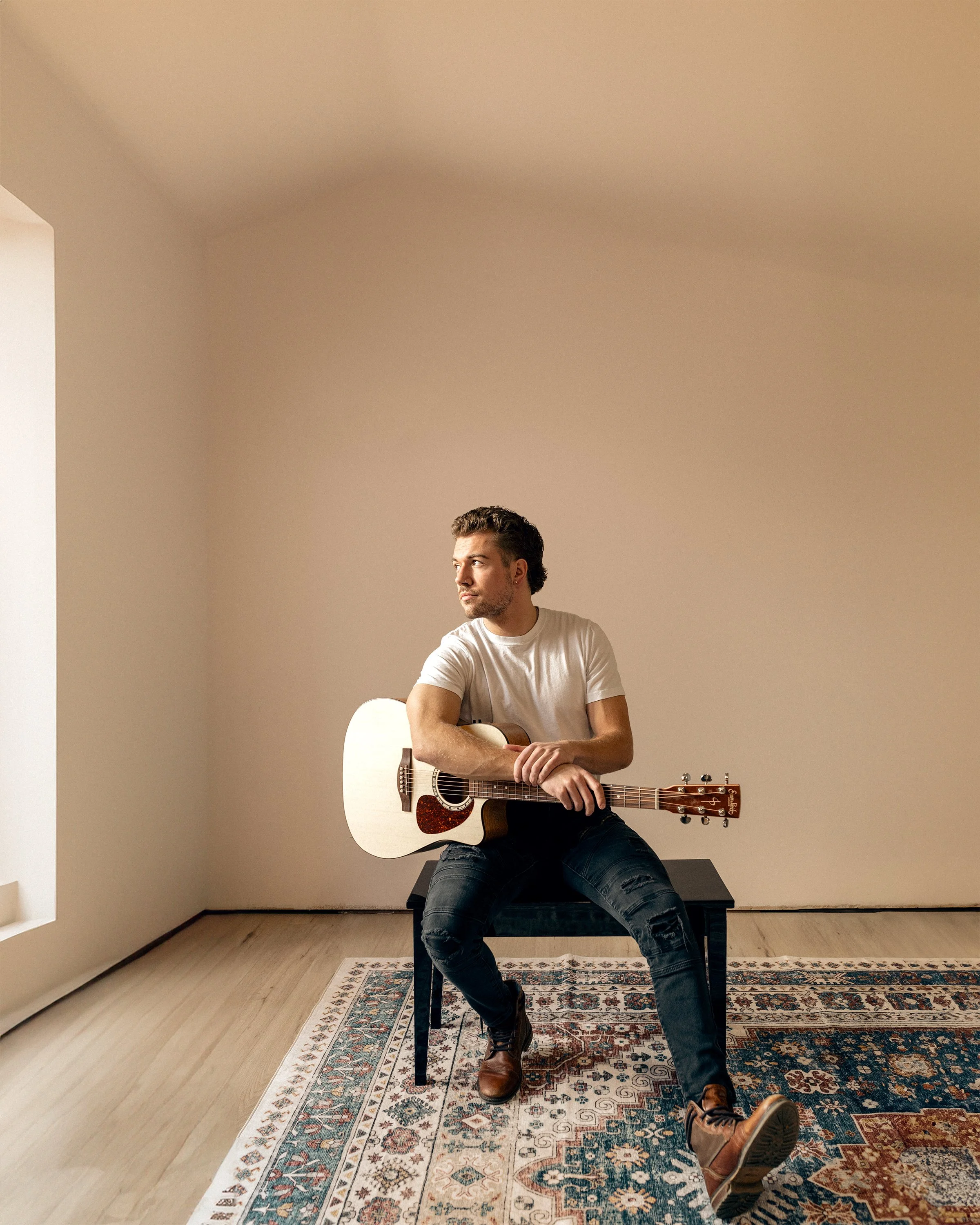 A young man with dark hair, wearing a white T-shirt and jeans, is sitting on a stool indoors, holding an acoustic guitar across his lap. He is looking off to the side with a thoughtful expression. The room has a neutral-colored wall, a patterned rug 