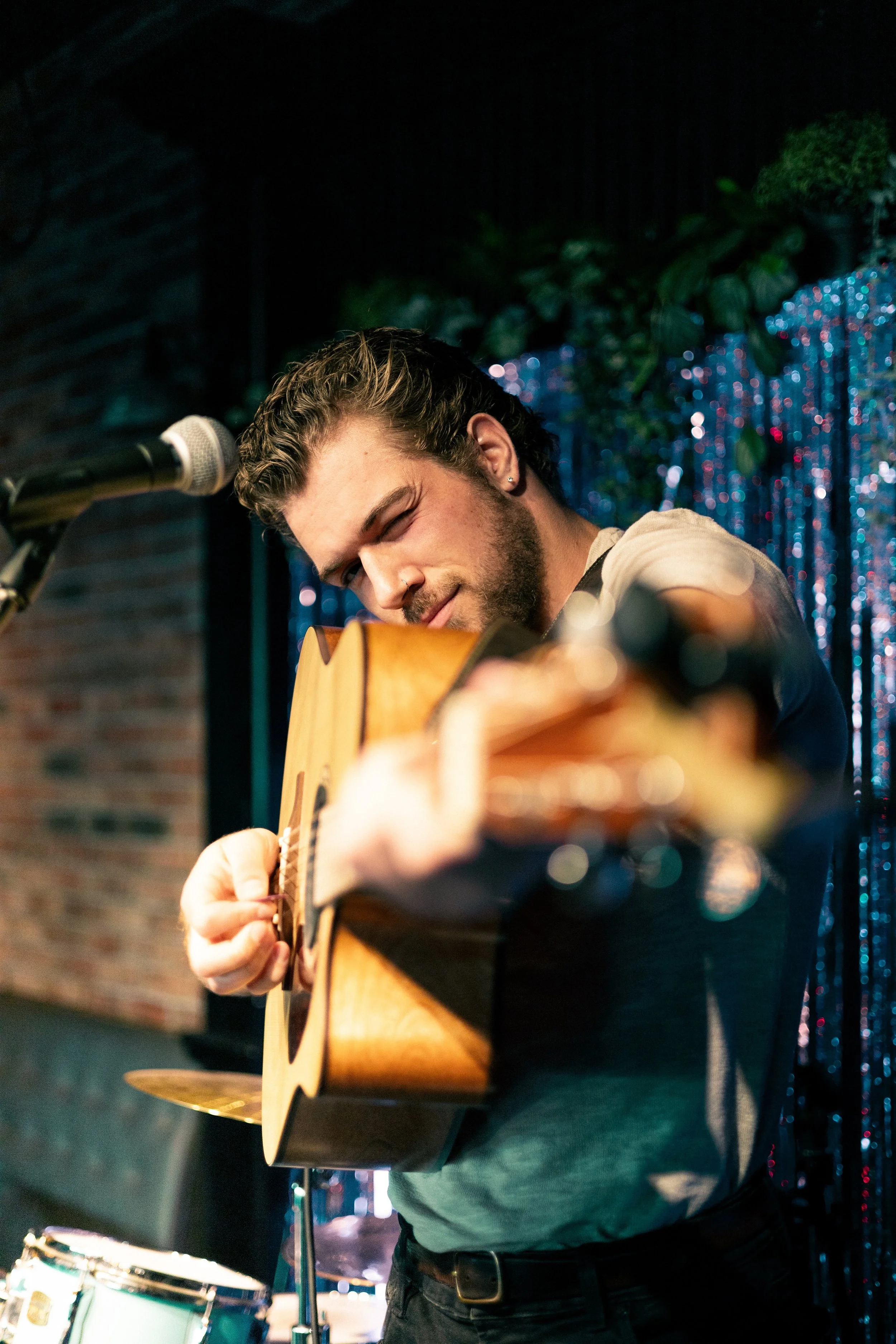 Matt Johner. A young man with styled hair and earrings playing an acoustic guitar on stage with a colorful, sparkly backdrop.