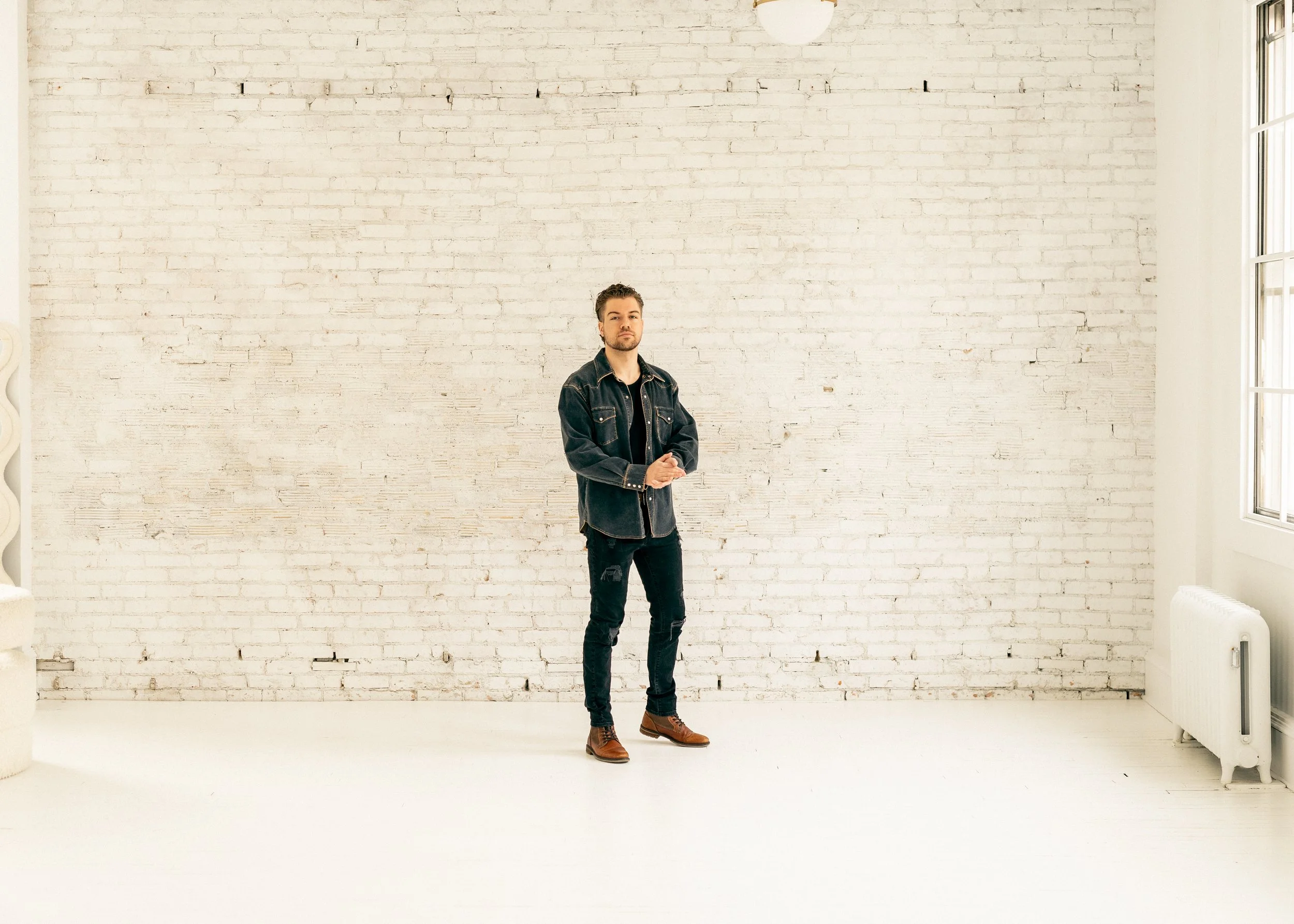 Matt Johner. A man standing with arms crossed in front of a white brick wall in a bright room with large windows.