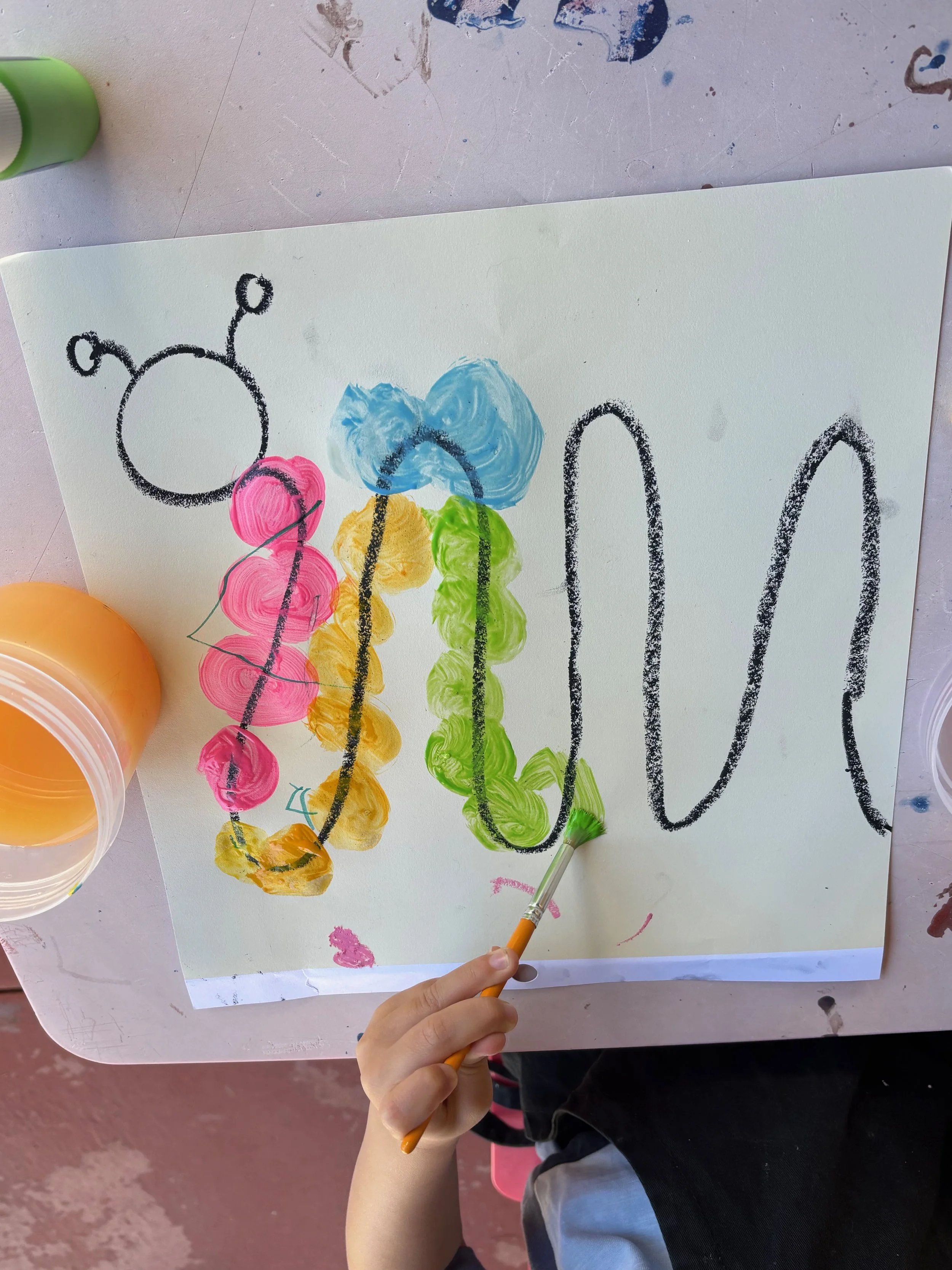 Child painting colorful circles and lines on a white sheet of paper with black marker and orange paintbrush at a pink table, surrounded by containers of orange and yellow paint.