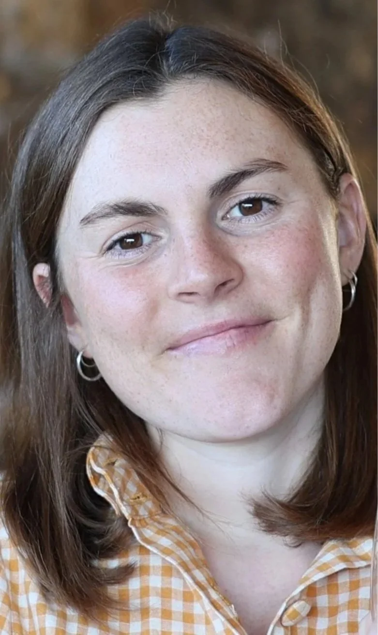 Close-up of a woman with medium-length brown hair, light skin, and brown eyes, wearing a yellow and white checkered shirt and silver hoop earrings.