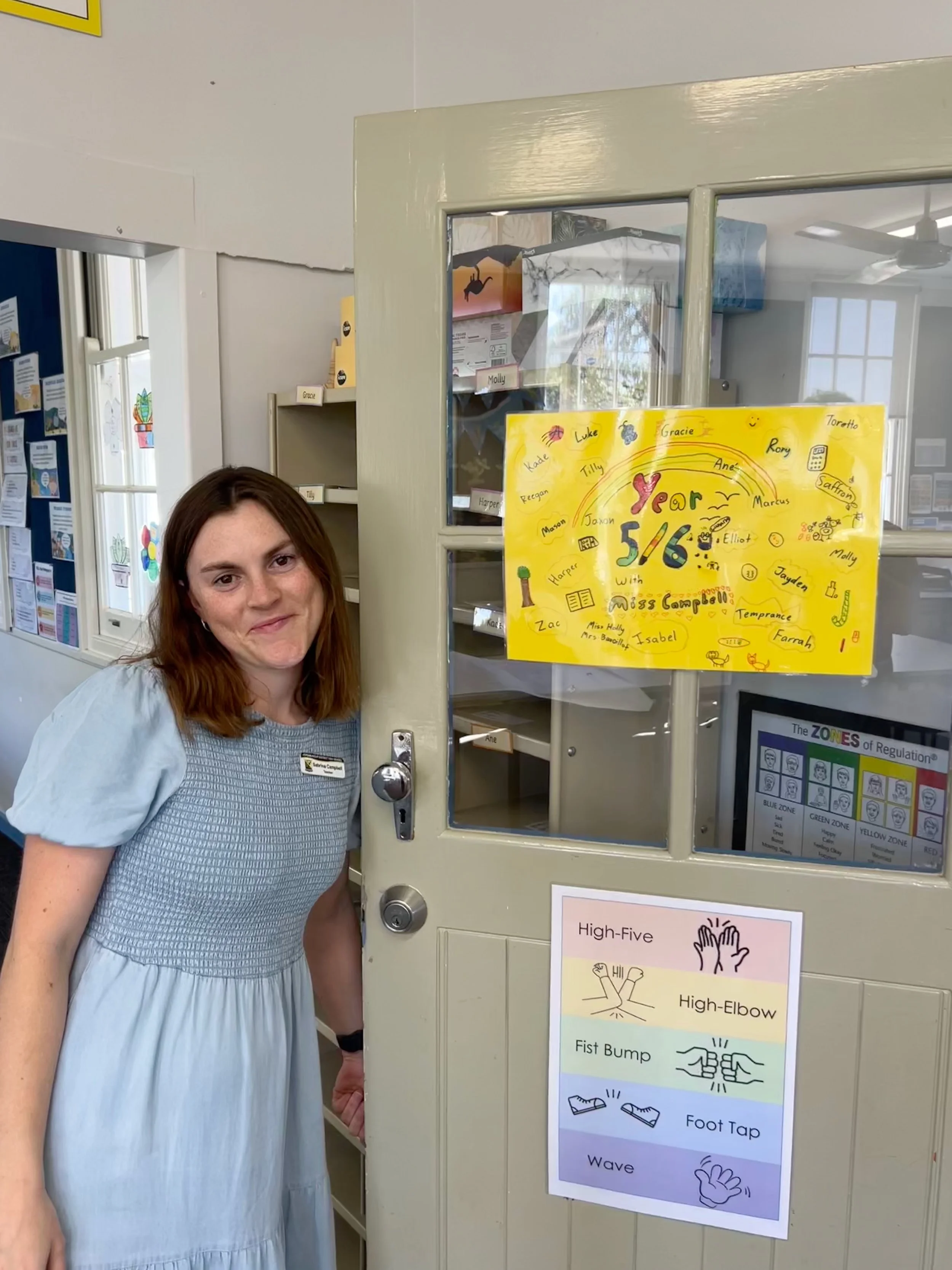 A woman with shoulder-length brown hair, wearing a light blue dress, stands next to an educational poster on a door. The poster features hand-drawn illustrations and text about various physical gestures such as high-five, high-elbow, fist bump, foot tap, and wave.