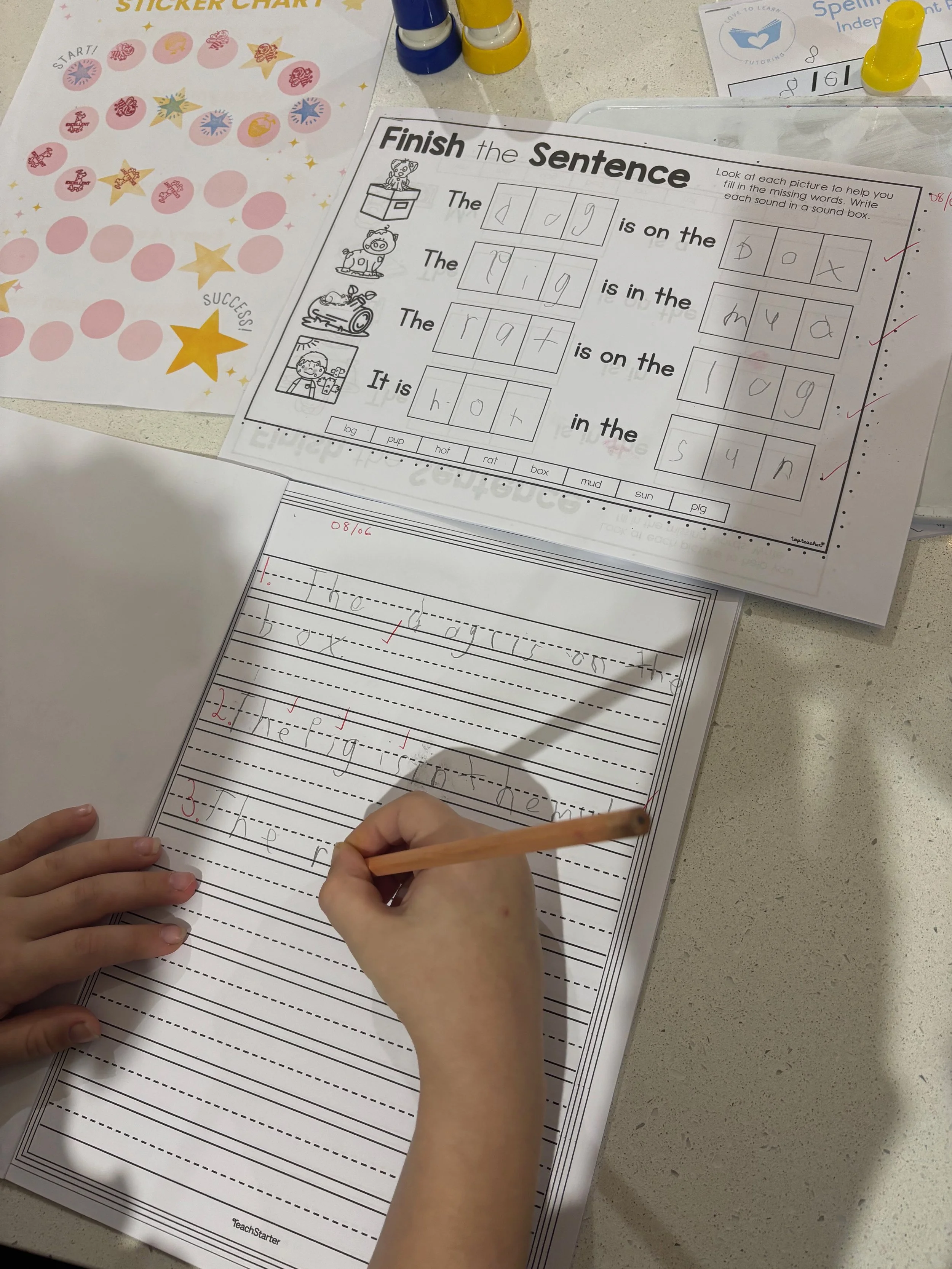 Close-up of a child's hands writing on a worksheet with a pencil, surrounded by other educational materials on a table, including a sticker chart and matching activity worksheet with pictures and words.