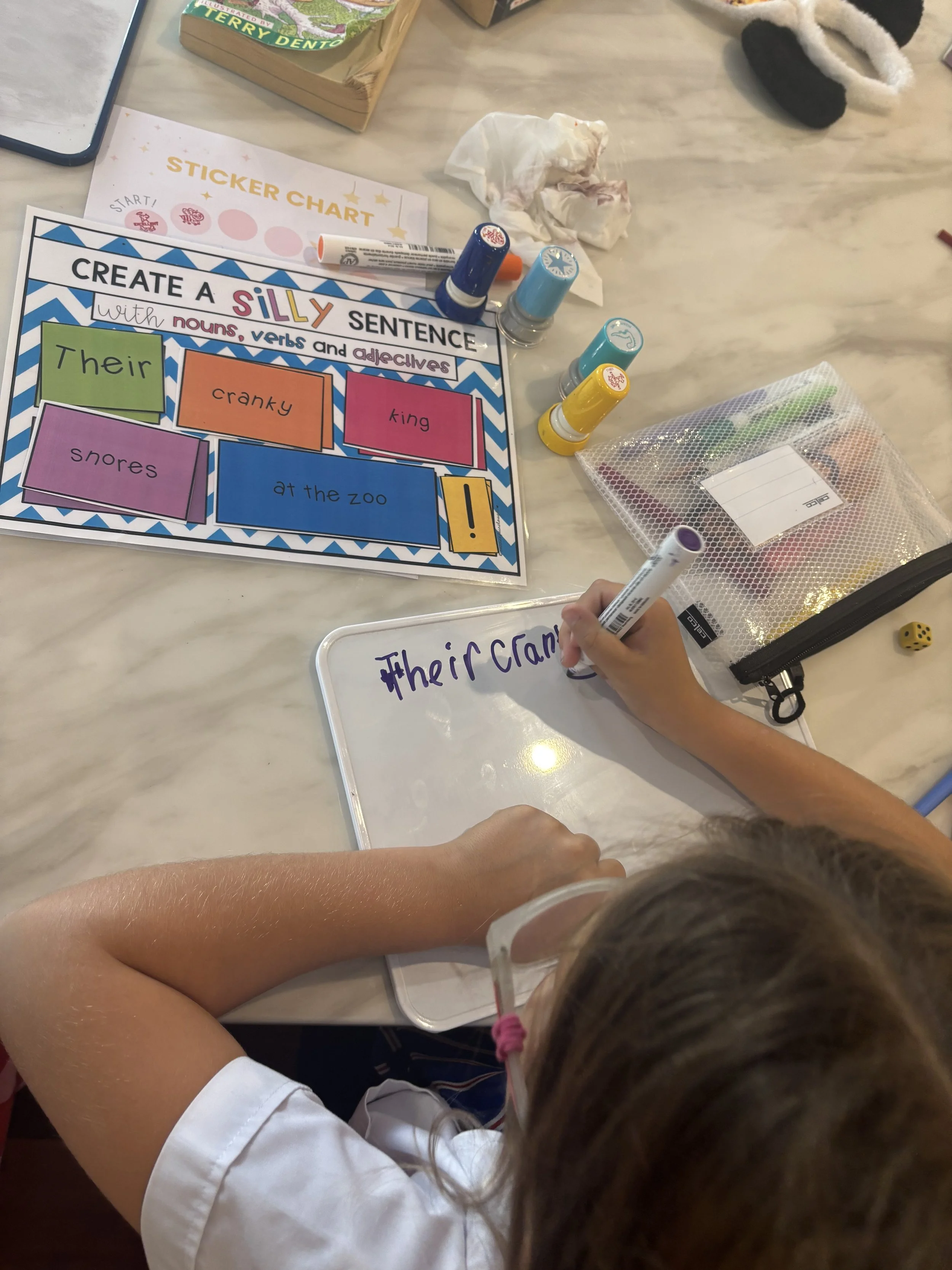 Children working on a colorful educational activity sheet titled 'Create a Silly Sentence' with a whiteboard and markers on a table.