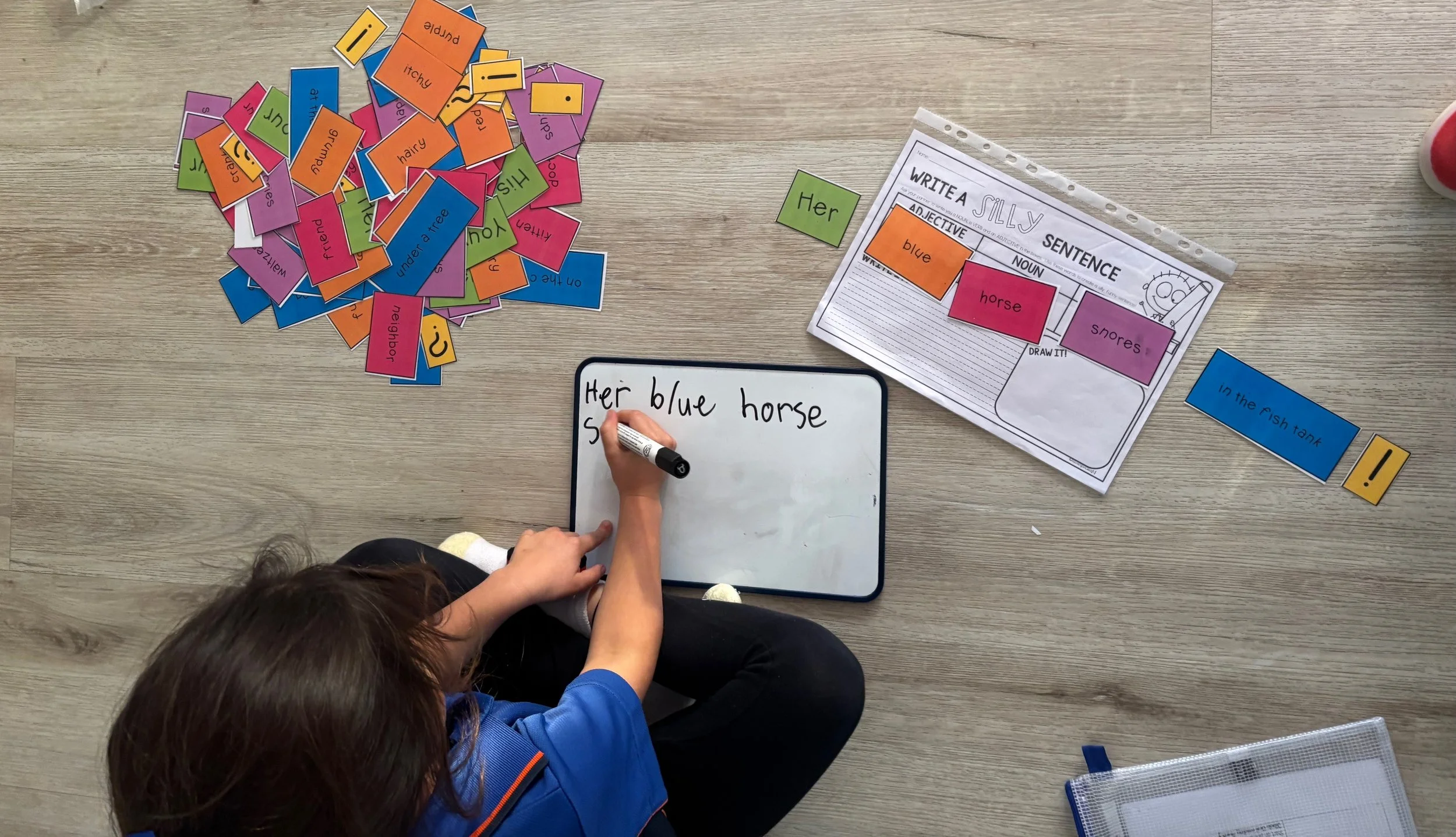 A child is writing on a small whiteboard with a black marker, with a activity sheet and colorful word cards scattered on a wooden floor.