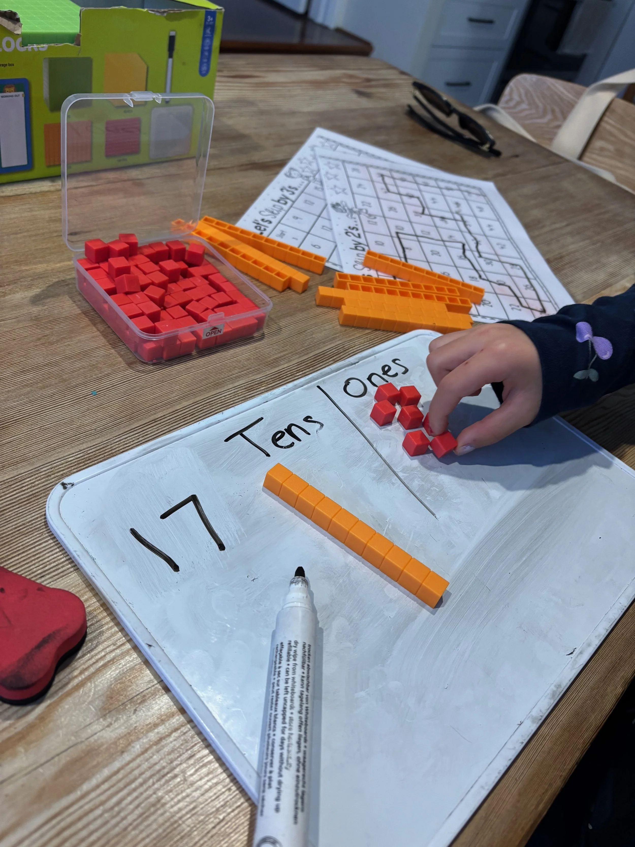 A child is sorting orange and red Connect 4 game pieces on a whiteboard labeled 'Tens' and 'Ones'. There is a plastic storage container filled with red game pieces, a game instruction sheet, and a dry erase marker on a wooden table.