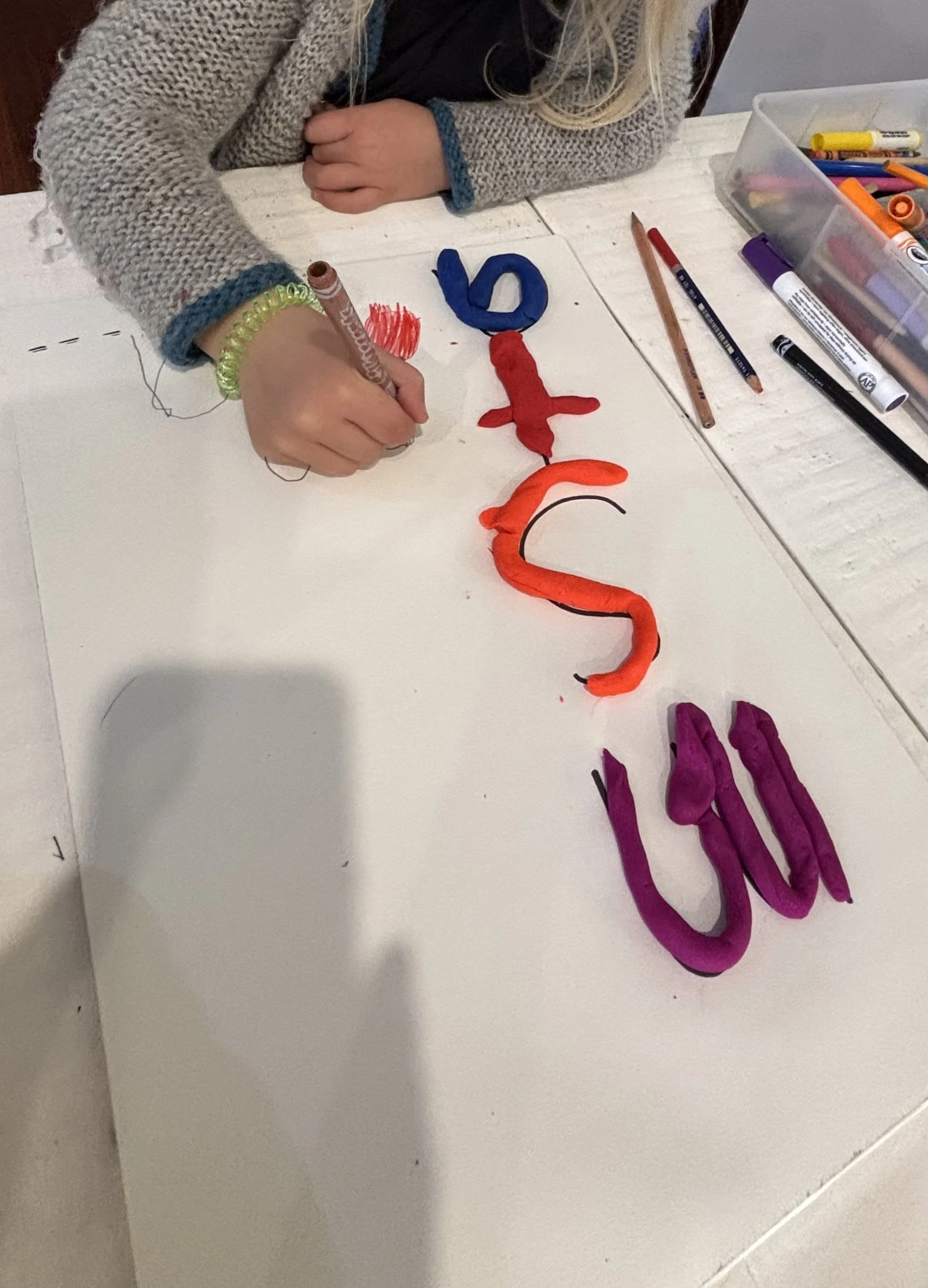 A child wearing a gray sweater with blue cuffs drawing on a white surface with a marker. The child has a green bracelet and a yellow bracelet on the left wrist. There are colorful drawings and clay sculptures of a fish, a snake, and the word "fun" on the surface. Office supplies such as markers, colored pencils, and scissors are organized in a plastic container nearby.