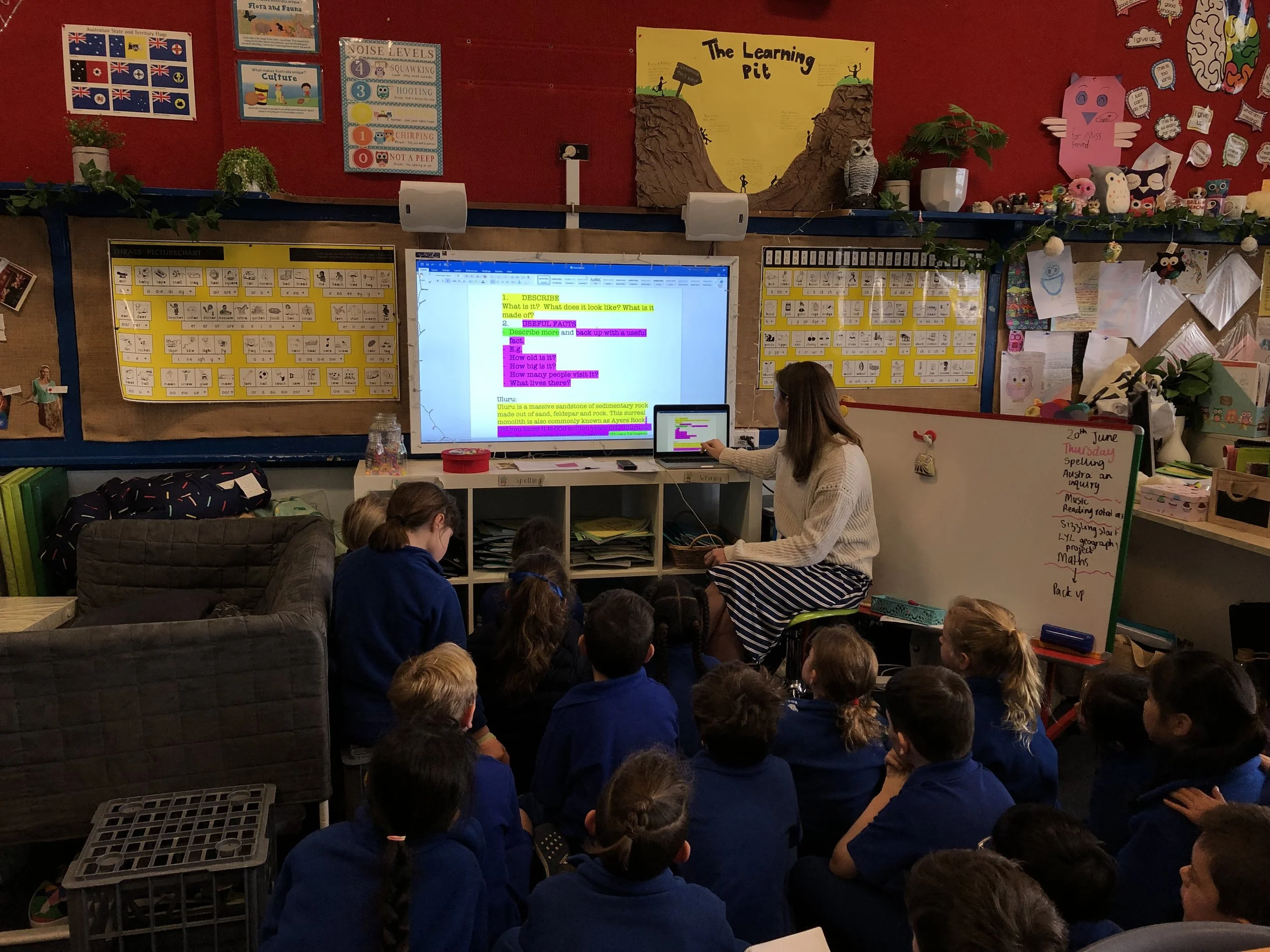 A classroom filled with young students sitting on the floor, facing a teacher who is seated at a desk. The classroom walls are decorated with educational posters, drawings, and colorful displays. There is a large screen behind the teacher displaying text. The classroom has a busy and vibrant atmosphere with various learning aids and decorations.