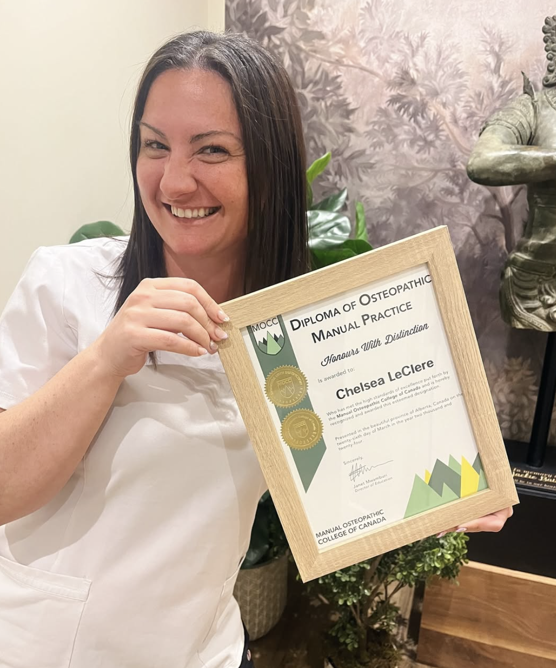 A woman smiling and holding a framed diploma of osteopathic manual practice award.