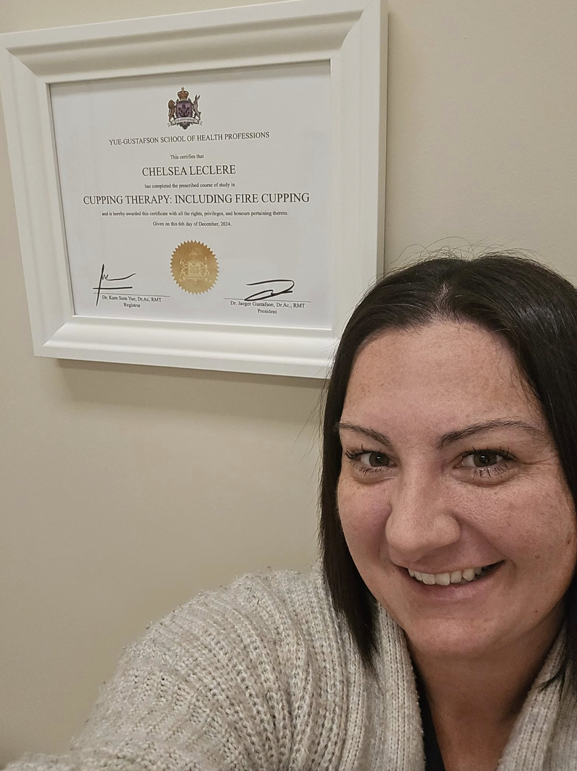 A woman taking a selfie in front of a framed certificate on the wall. The certificate is from the Yue-Gustafson School of Health Professions for completing a course in cupping therapy, including fire cupping, awarded on December 6, 2024.