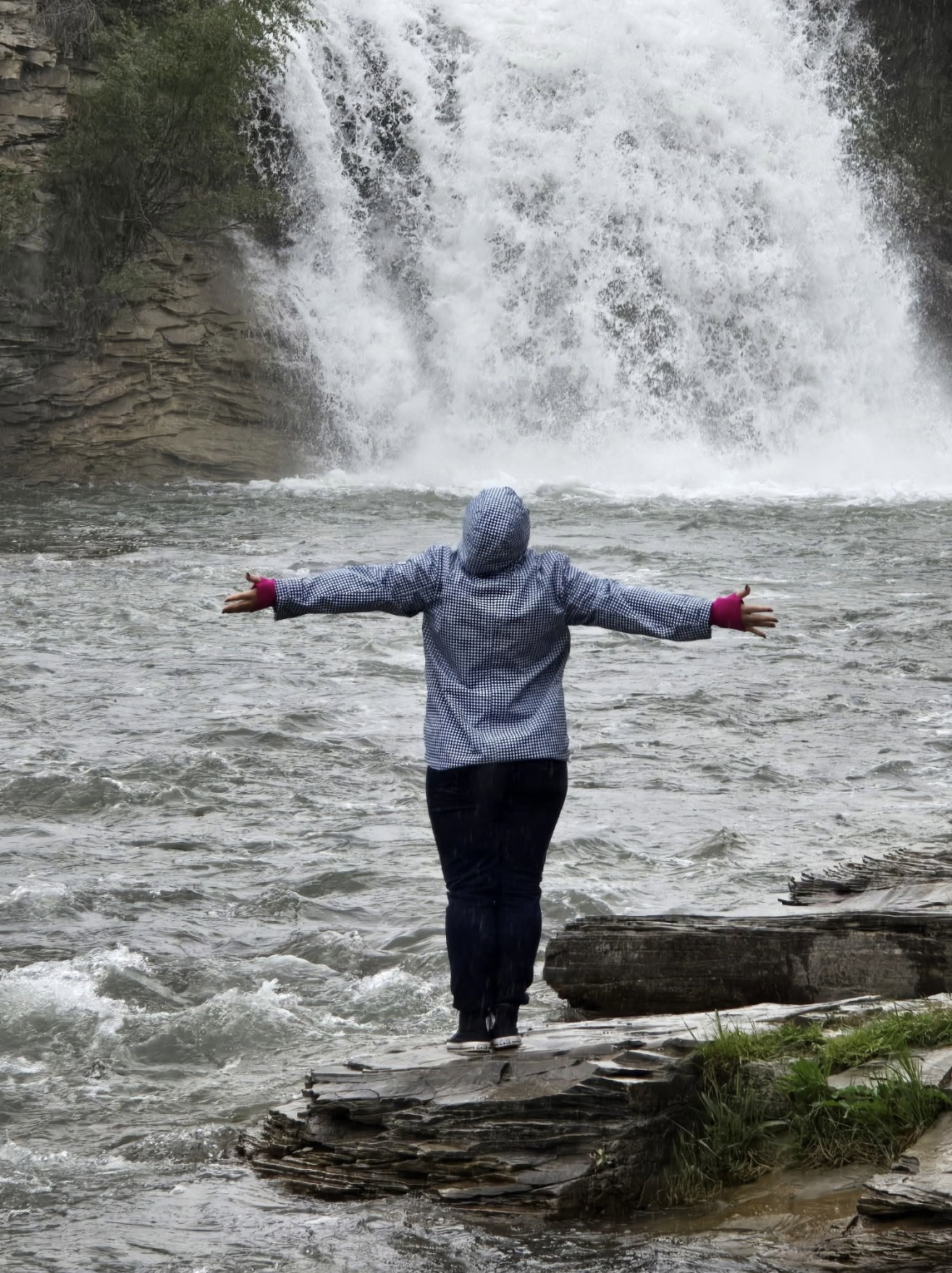 Person standing on rocks near a waterfall with arms outstretched