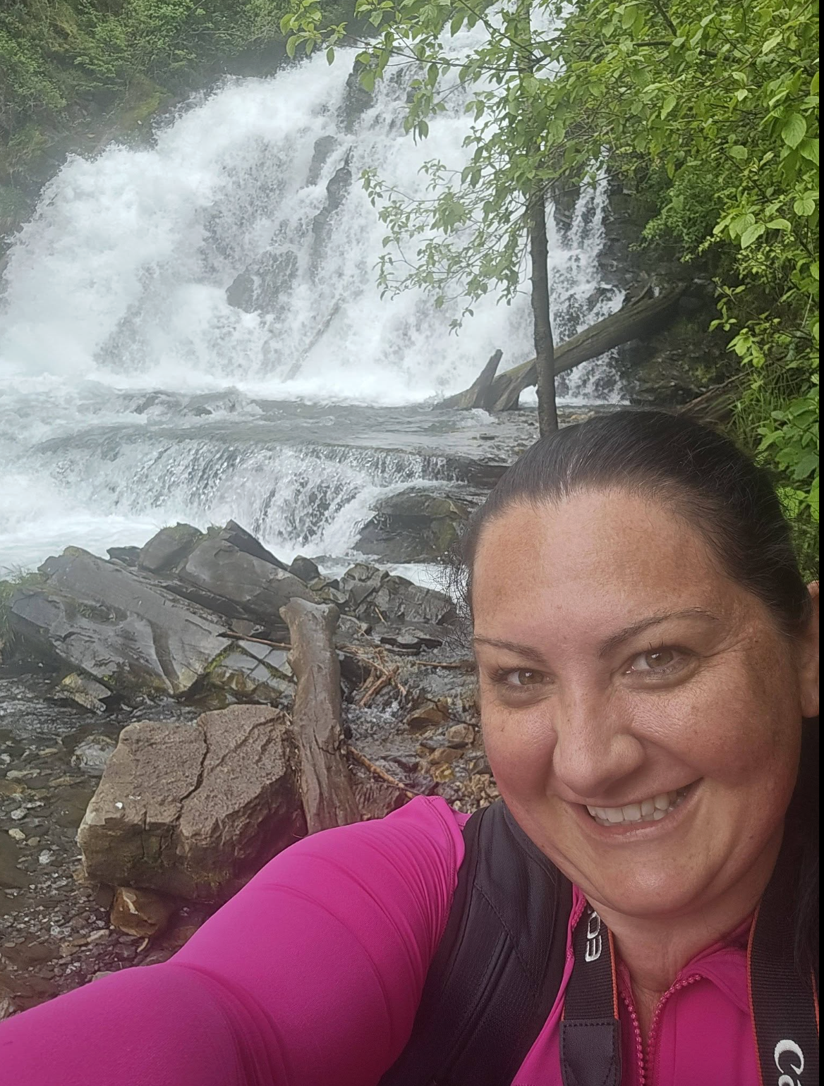 A woman smiling and taking a selfie in front of a waterfall surrounded by green trees and rocks.