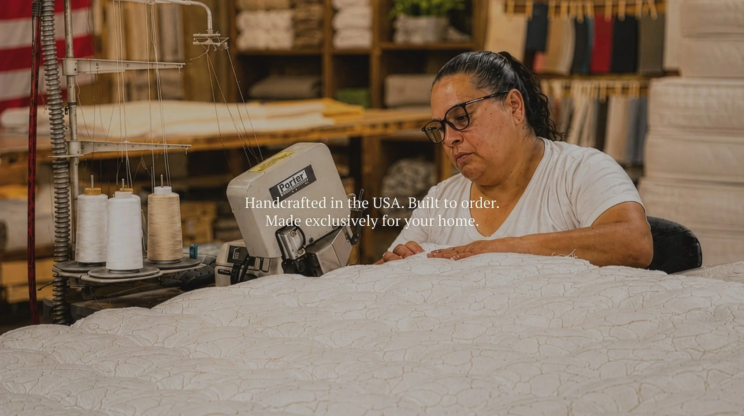 A woman with dark hair and glasses sewing on a machine in a workshop with shelves of fabric in the background.