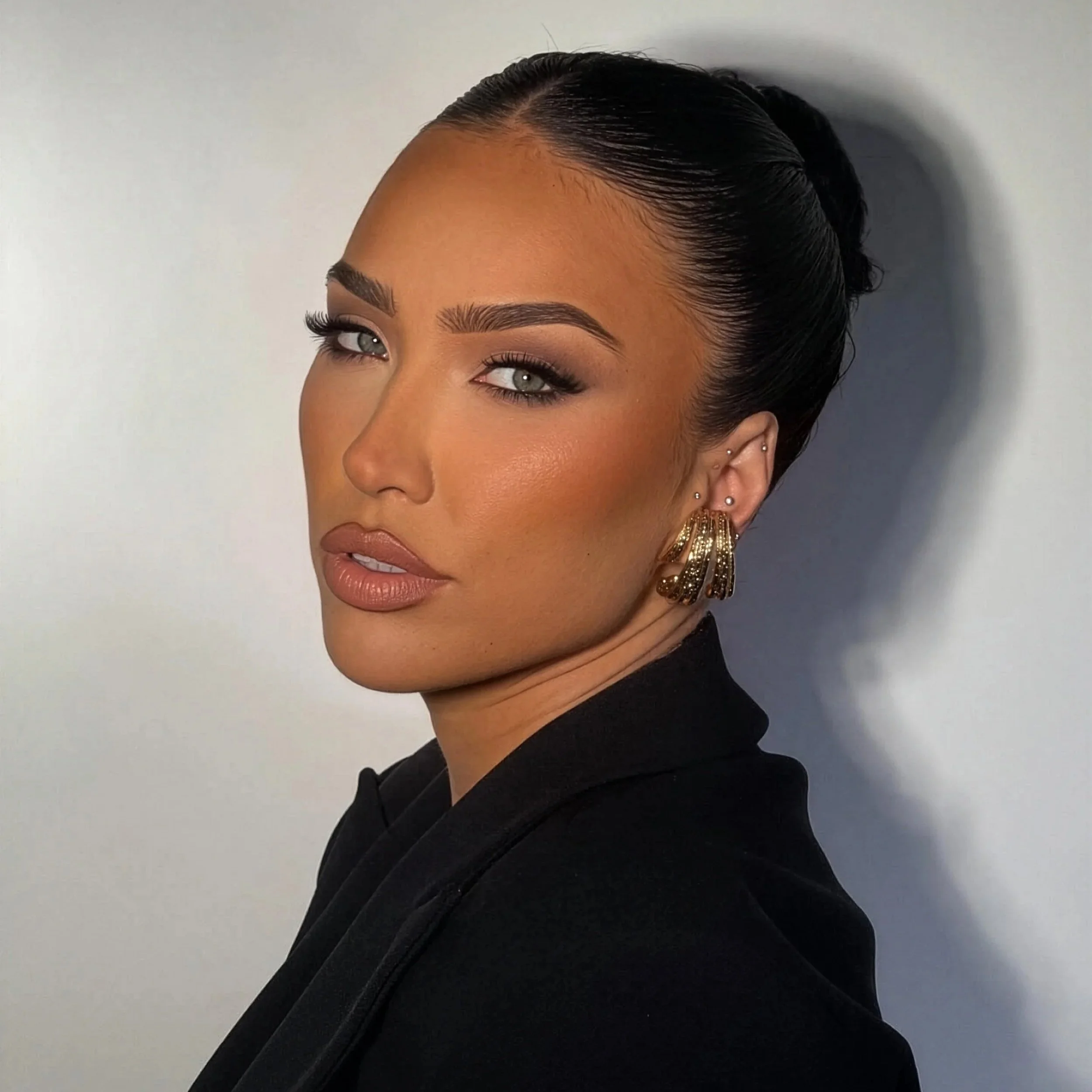 Close-up of a woman with dark hair styled in a sleek, slicked-back bun, wearing gold earrings, with makeup emphasizing her eyes and eyebrows, against a neutral background.