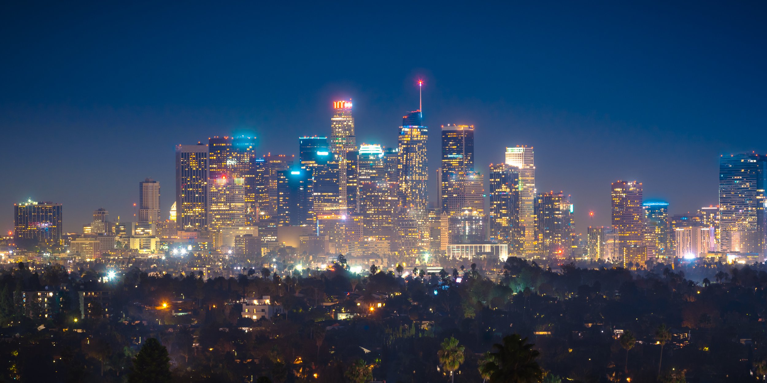 Nighttime view of Los Angeles city skyline with tall illuminated skyscrapers and a dark blue sky.