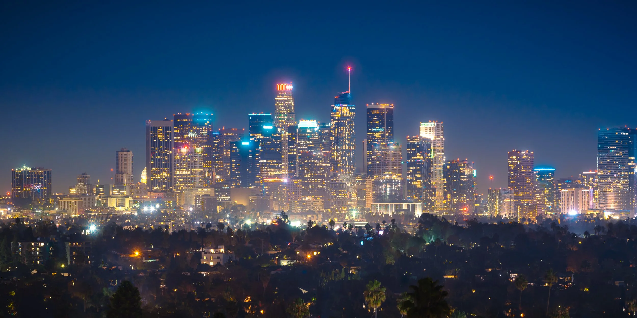 Nighttime view of a city skyline with tall illuminated skyscrapers and a dark blue sky.