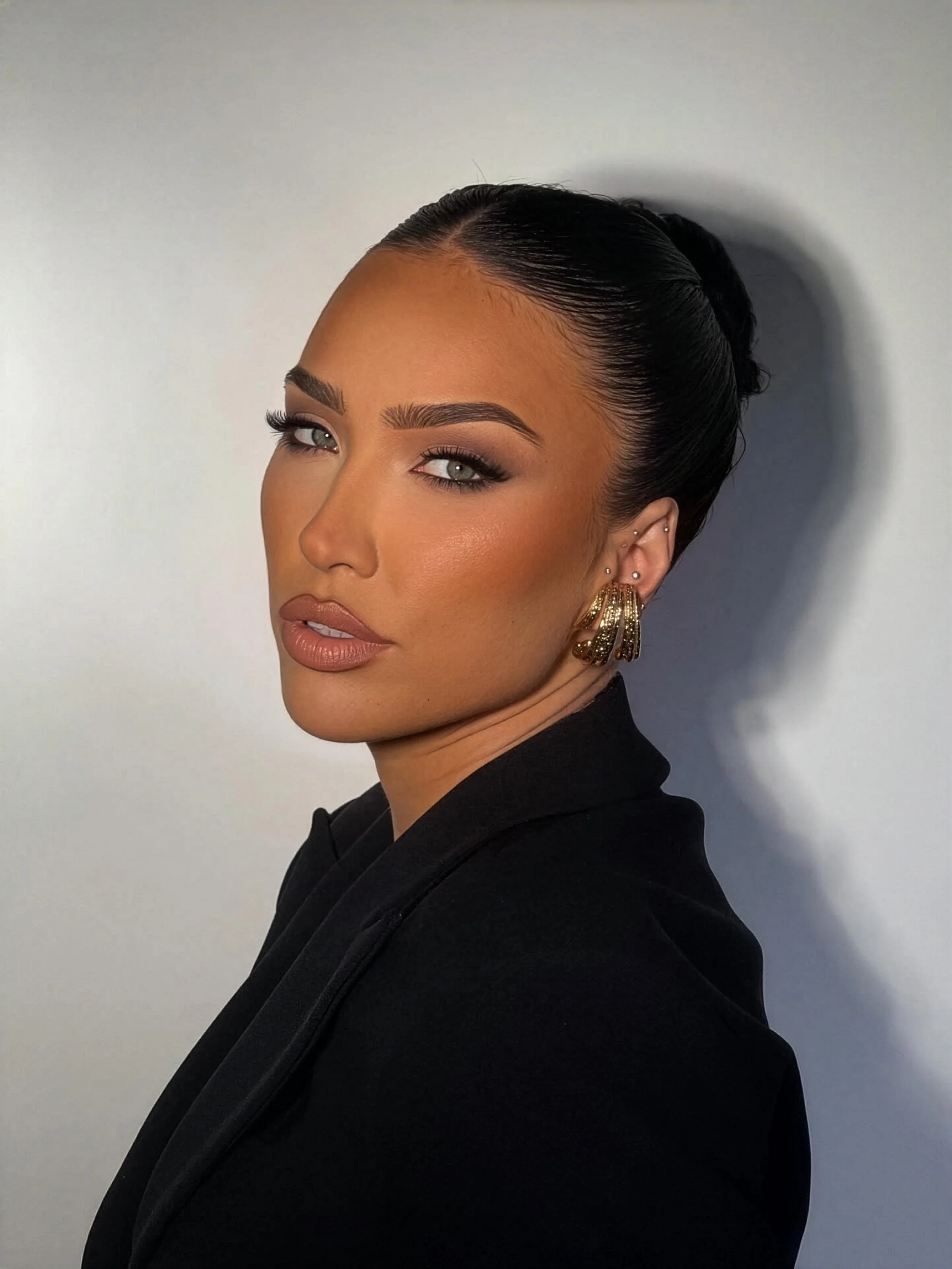 Close-up portrait of a woman with sleek black hair styled in an elegant updo, wearing gold earrings and makeup with defined eyebrows, long eyelashes, and neutral lipstick, against a plain light-colored background.