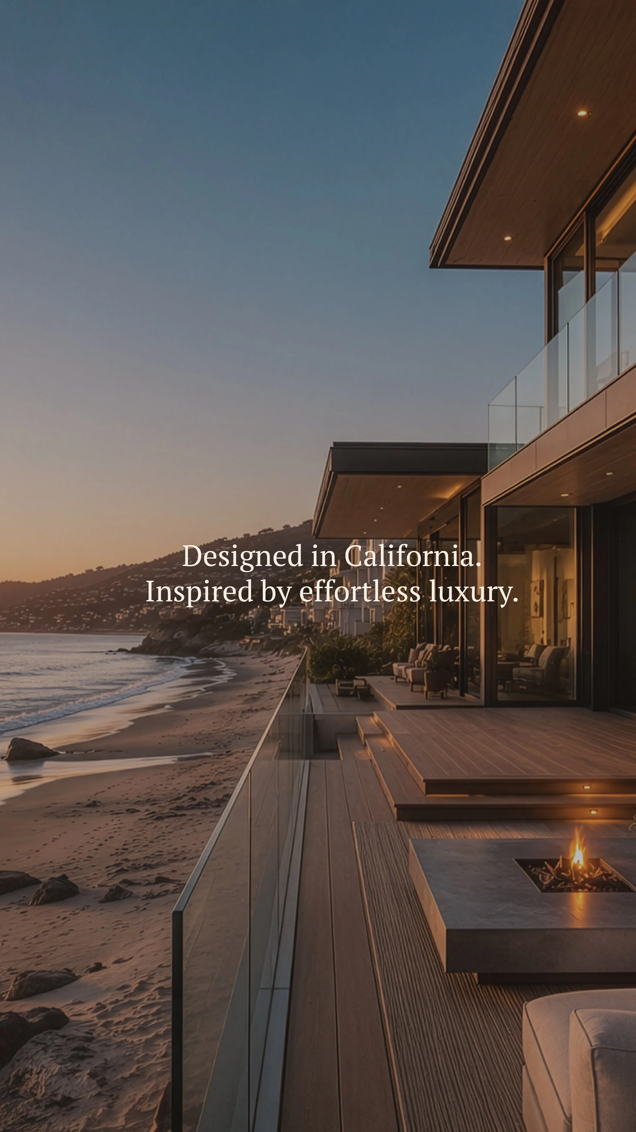 Modern house with large glass windows overlooking a beach at sunset, with outdoor seating and a fire pit.